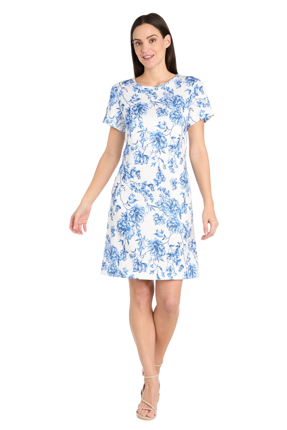 A woman stands facing forward against a plain white background, wearing the R&M Richards 6674P Petite Print Party Dress—short-sleeved, knee-length, white with a blue floral pattern—paired with beige strappy heels.
