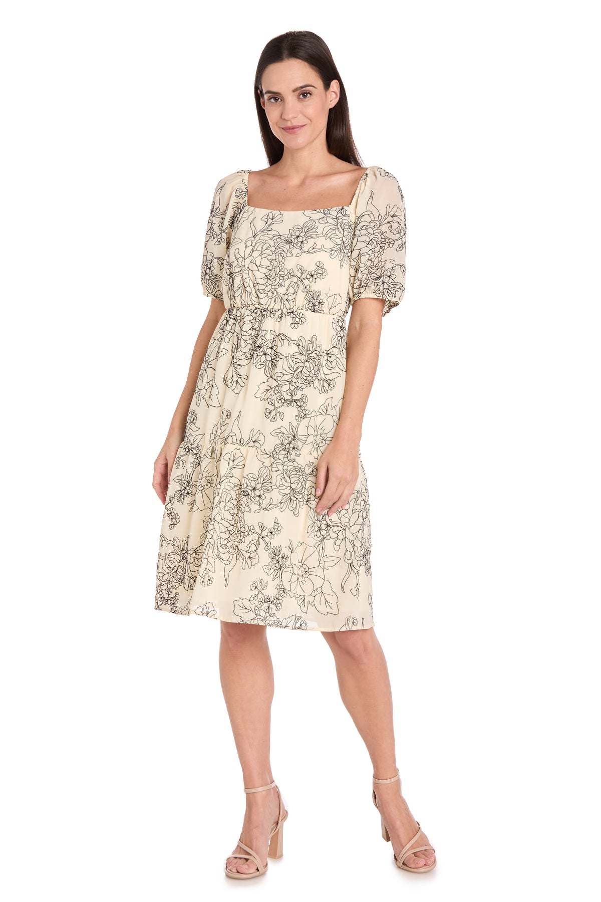 A woman wears the Petite Knee-Length Party Dress: R&M Richards 6688P, a cream short-sleeve floral dress with black patterns and beige heeled sandals, standing against a white background.