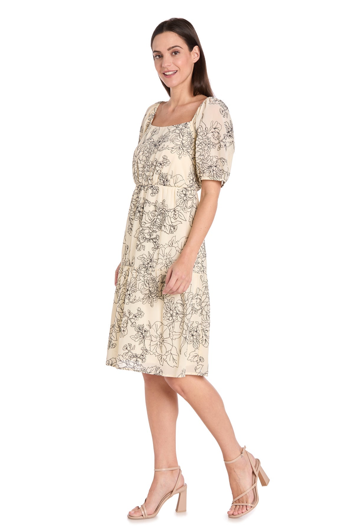 Woman in the Petite Knee-Length Party Dress: R&M Richards 6688P—light beige, short sleeves, black floral print, puffed sleeves—paired with nude high-heeled sandals, standing against a white background.