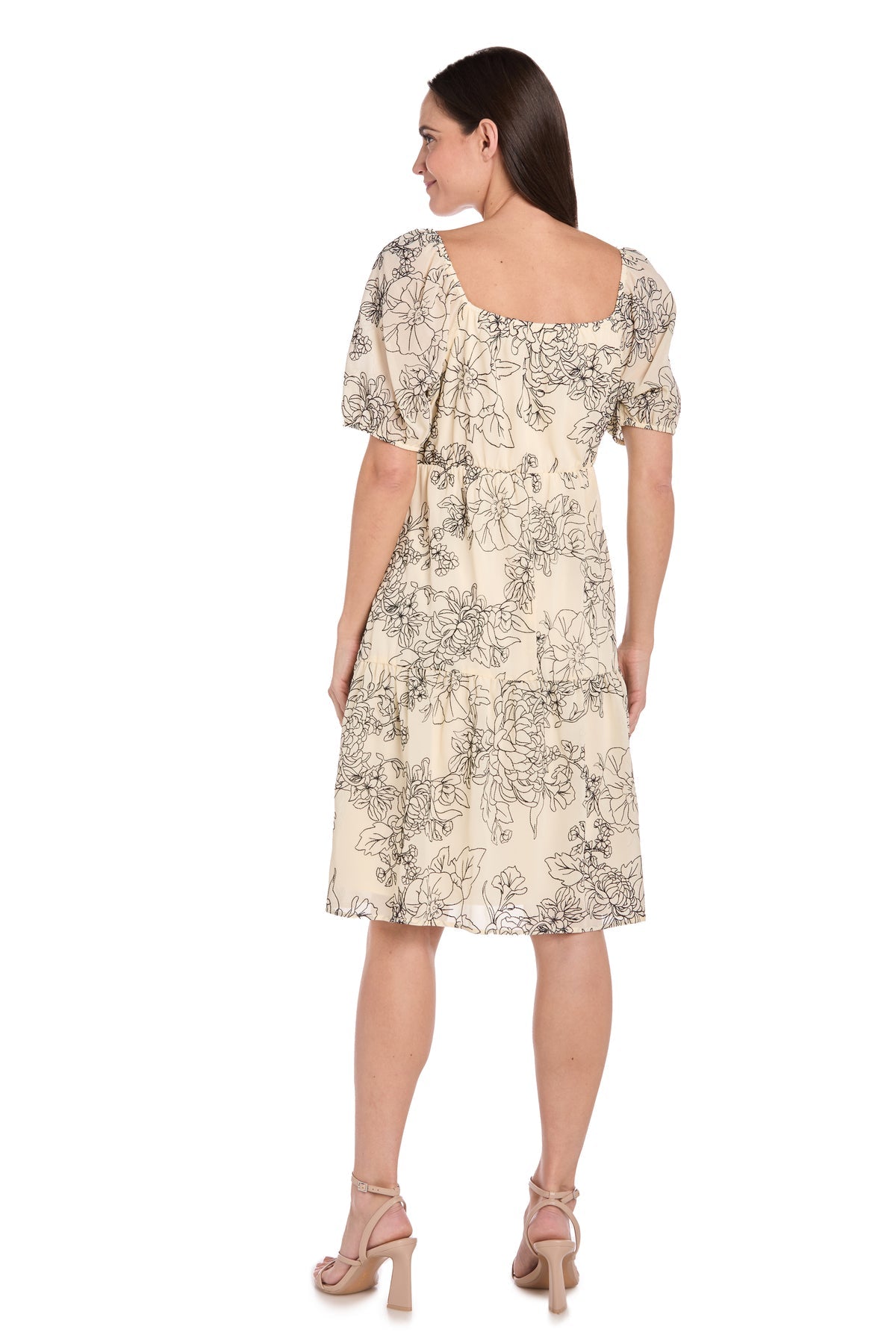 A woman faces away from the camera wearing the R&M Richards 6688P petite knee-length party dress, featuring a light-colored floral print, short puff sleeves, and a square neckline.