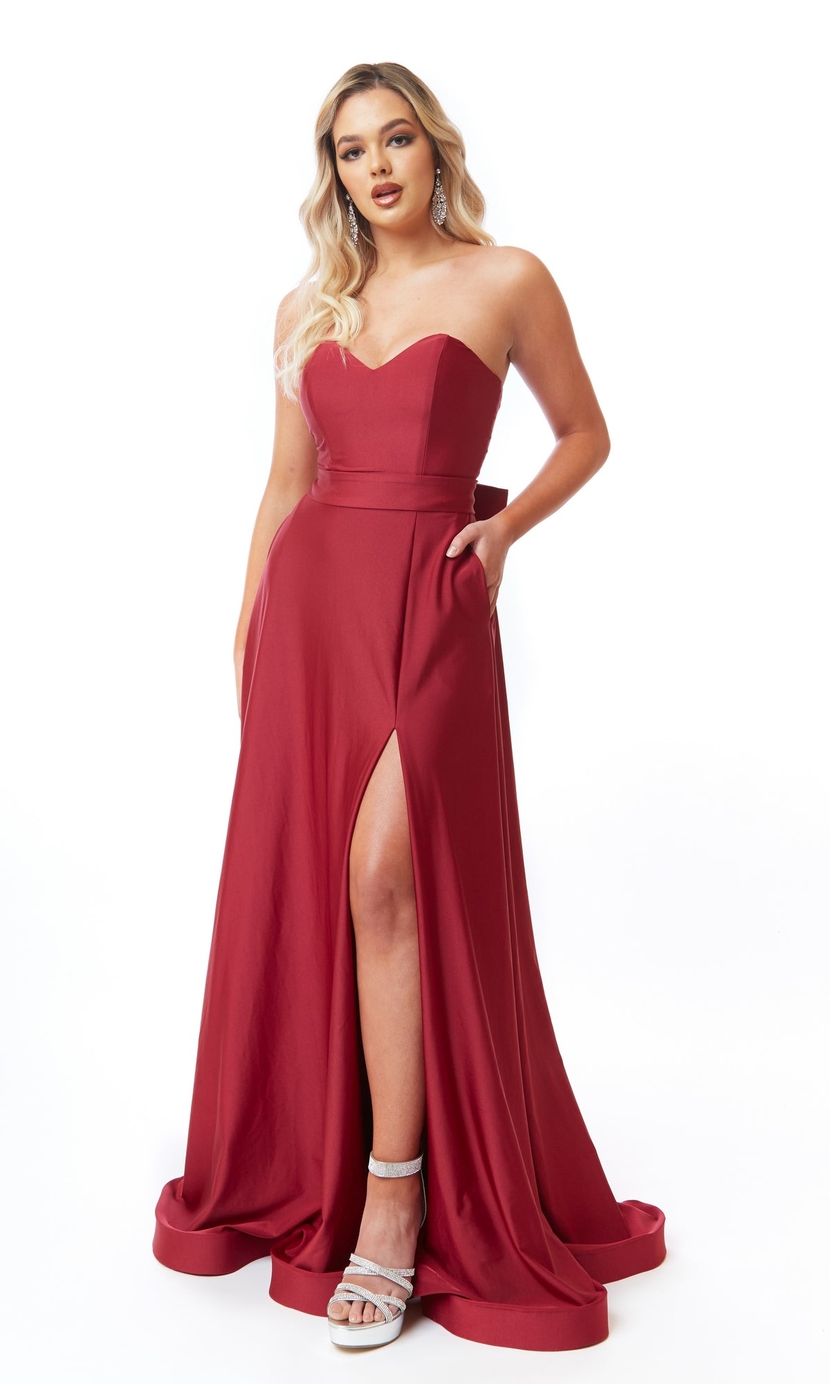 A woman in an Atria 6705H Strapless Sweetheart Long Prom Dress—a floor-length red gown with a thigh-high slit and pockets—pairs it with silver high-heeled sandals, standing against a white background.