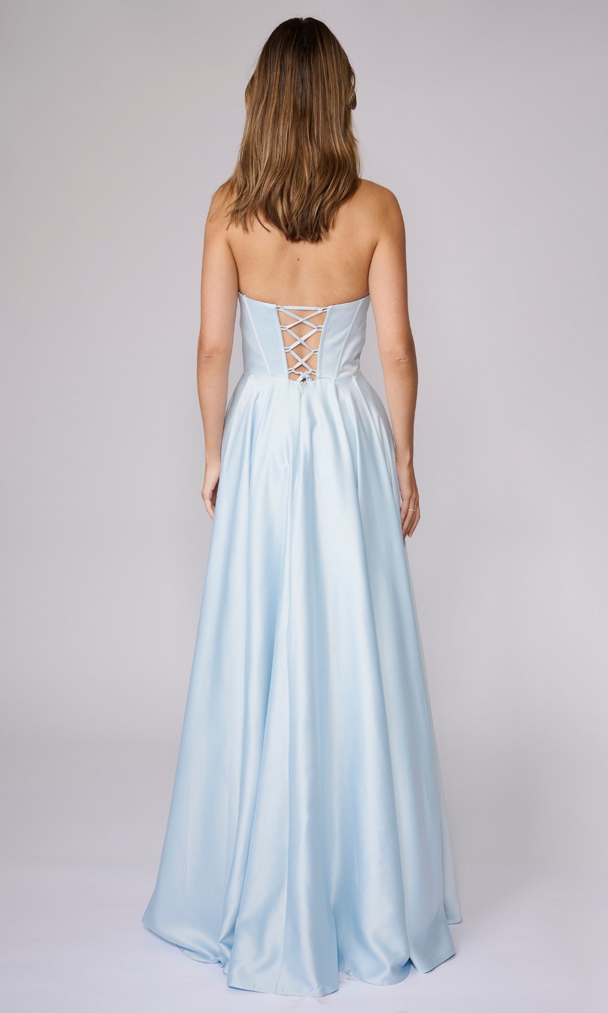 A woman stands with her back to the camera in a strapless light blue Nina Canacci 6718 long formal dress featuring an A-line silhouette and lace-up back.