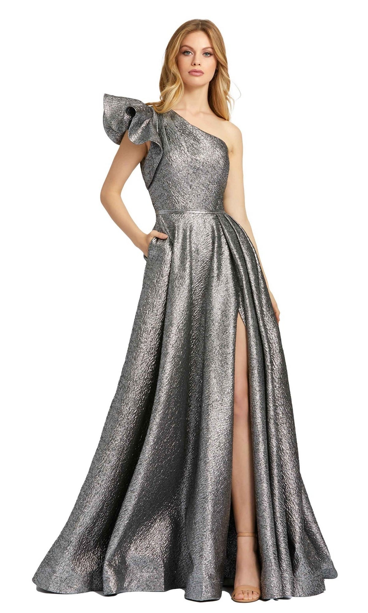 A woman stands elegantly against a white background wearing the Mac Duggal 67297 Metallic Long Prom Ball Gown, featuring a one-shoulder design, ruffled sleeve, high slit, and side pockets.