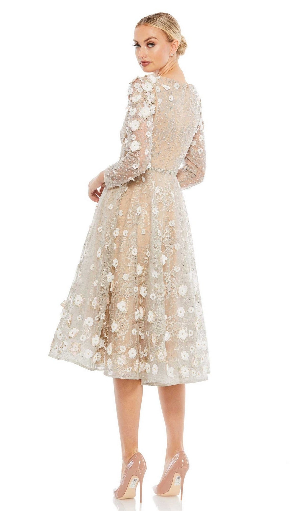 A woman in the Short Wedding Guest Dress 67387 by Mac Duggal—knee-length, sheer beige with floral embroidery and long sleeves—pairs it with nude heels, standing facing away and glancing over her shoulder.