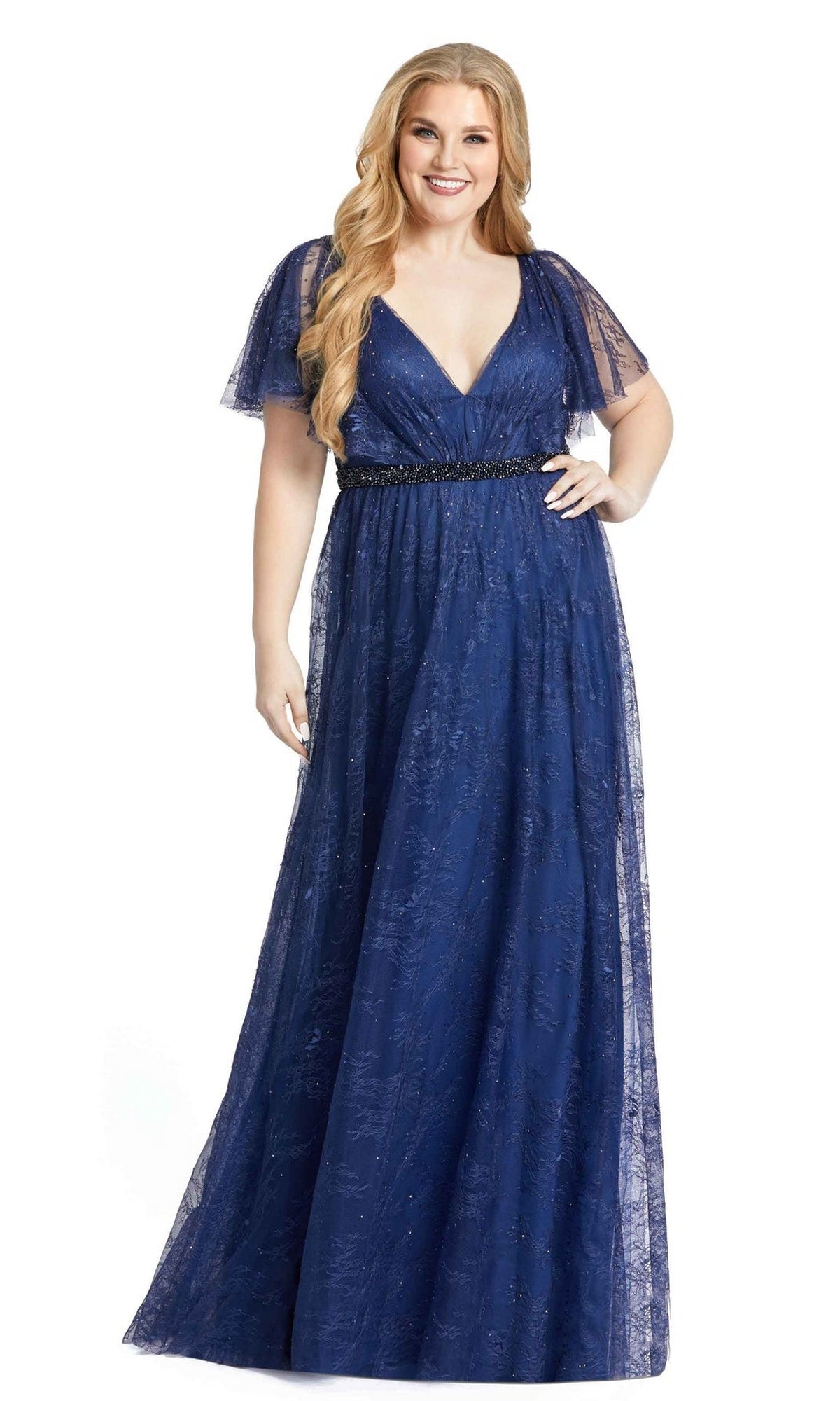 A woman in the Mac Duggal Long Plus-Size Prom Dress 67435, featuring a floor-length deep blue design with V-neckline, flutter sleeves, and a beaded waistband, stands smiling.