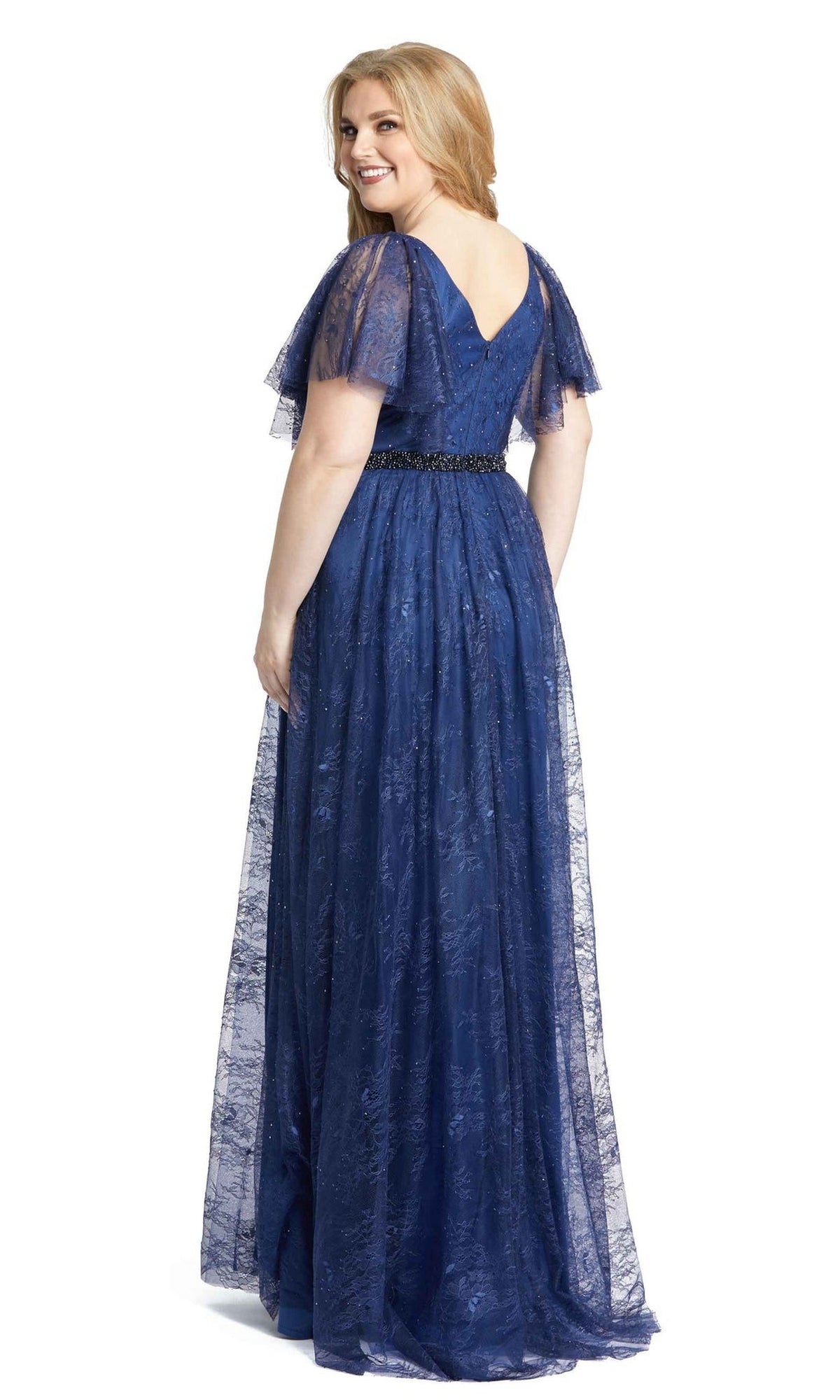 A woman in the Long Plus-Size Prom Dress 67435 by Mac Duggal, featuring a beaded waistband and lace A-line design, stands floor-length and turns to smile at the camera.