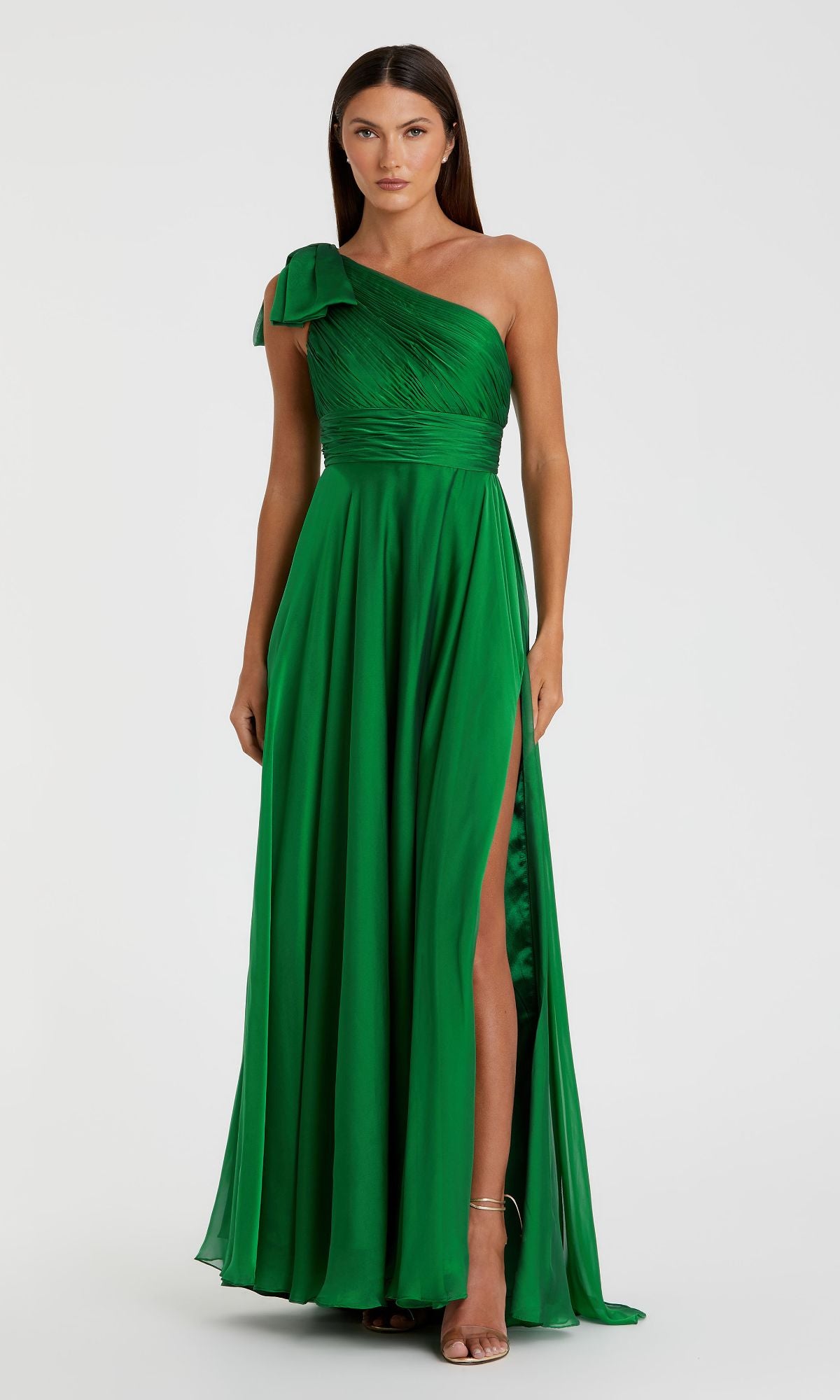 A woman wears the Mac Duggal 67476, a long one-shoulder green prom dress with a thigh-high slit, standing against a plain light background.