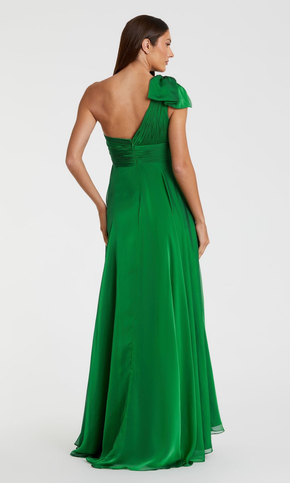 A woman faces away, wearing the Mac Duggal 67476 long one-shoulder prom dress—a floor-length green gown with a bow detail, highlighting the elegant back design of this stunning formal outfit.