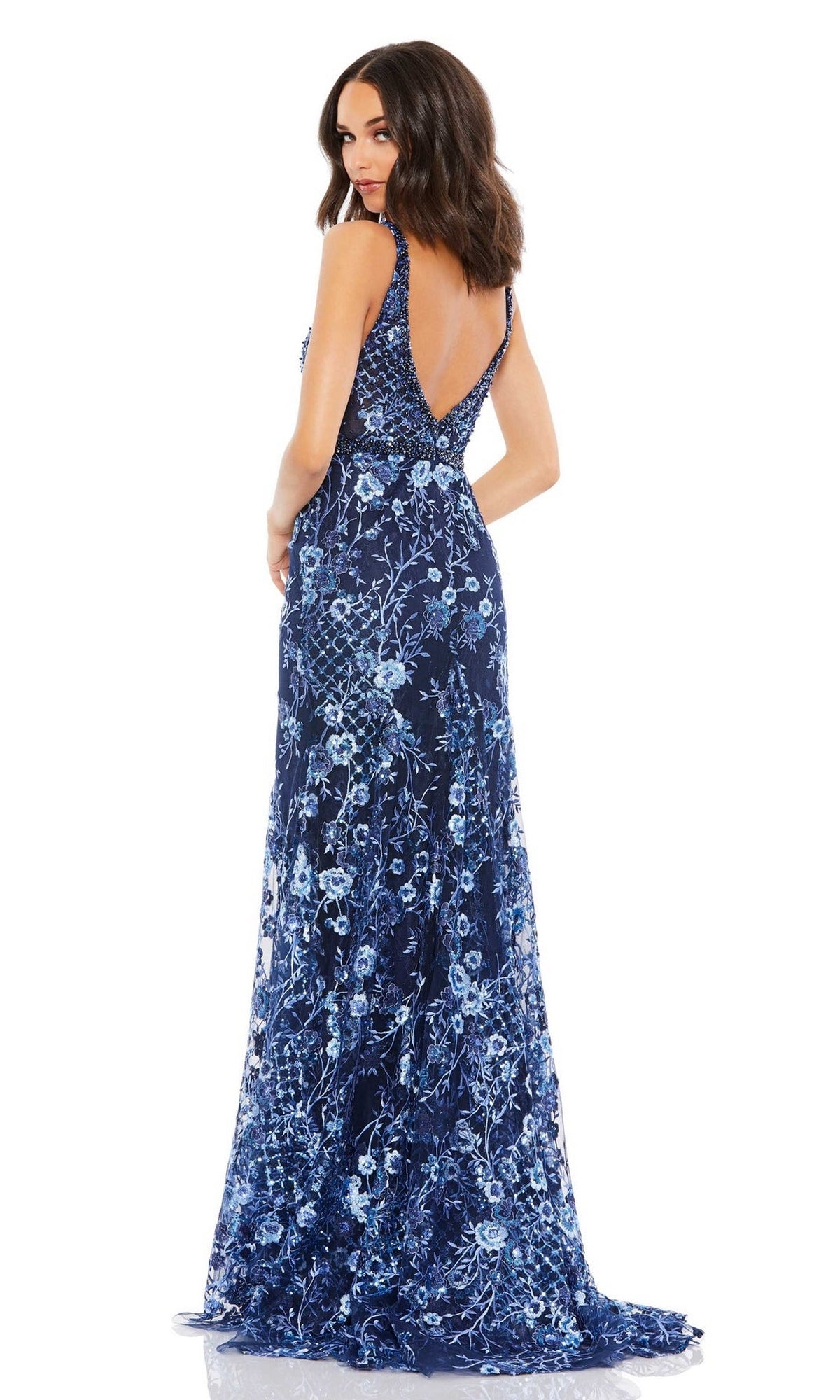 A woman stands facing away, wearing the Mac Duggal 67481 Long Floral-Print Formal Dress featuring a sleeveless design, deep blue floral prints, and a low V-back.