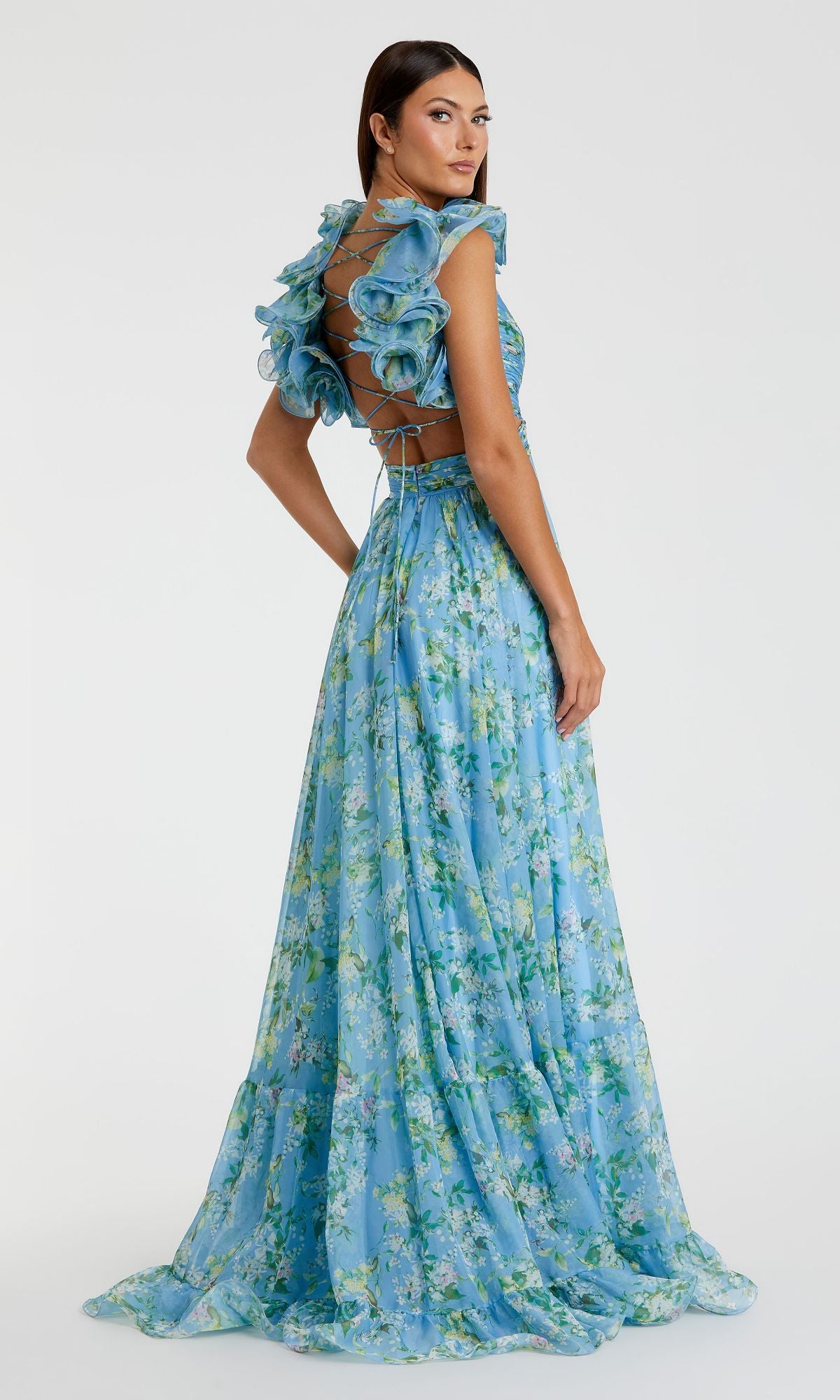 A woman stands looking over her shoulder in the Mac Duggal 67803 Ruffled Long Print Formal Dress, featuring a blue floral pattern, ruffled sleeves, and a lace-up back.