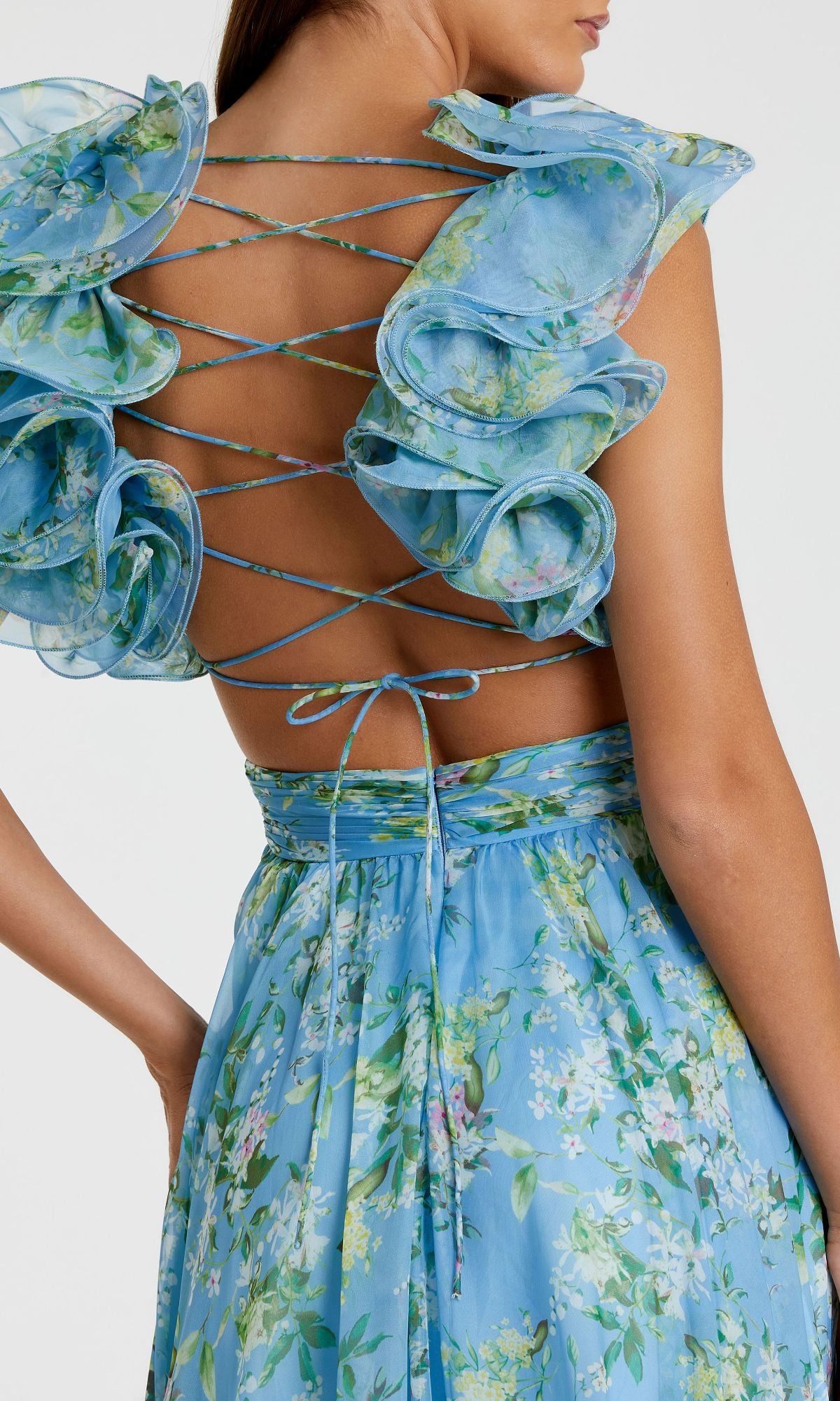 A woman wears the Ruffled Long Print Formal Dress: Mac Duggal 67803, featuring a blue floral print, ruffled details, an open back, and crisscrossed laces tied at the lower back.