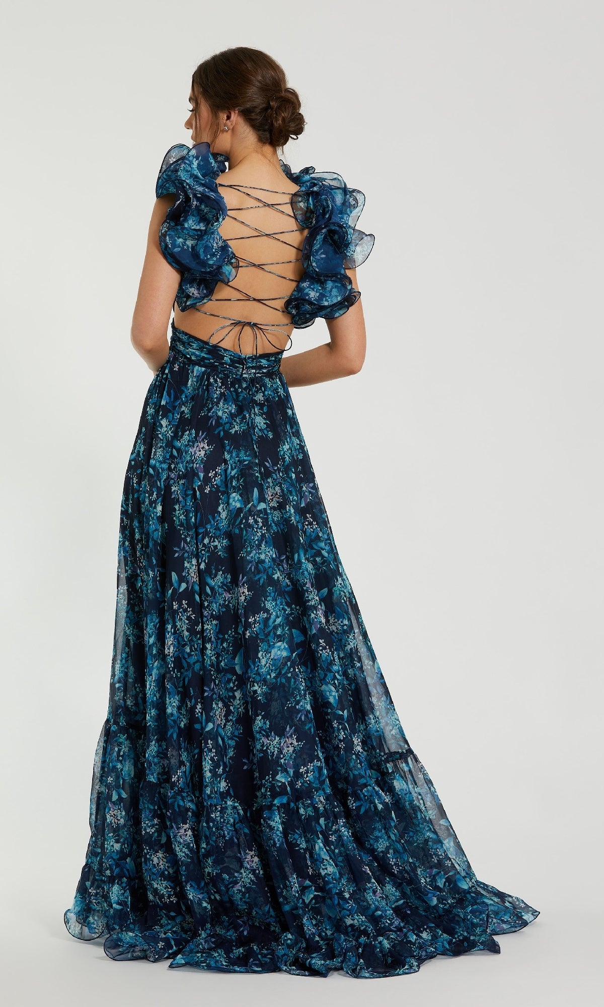 A woman stands facing away from the camera in the Mac Duggal 67803 Ruffled Long Print Formal Dress, a dark blue floral gown featuring ruffled sleeves and an open lace-up back, set against a plain background.