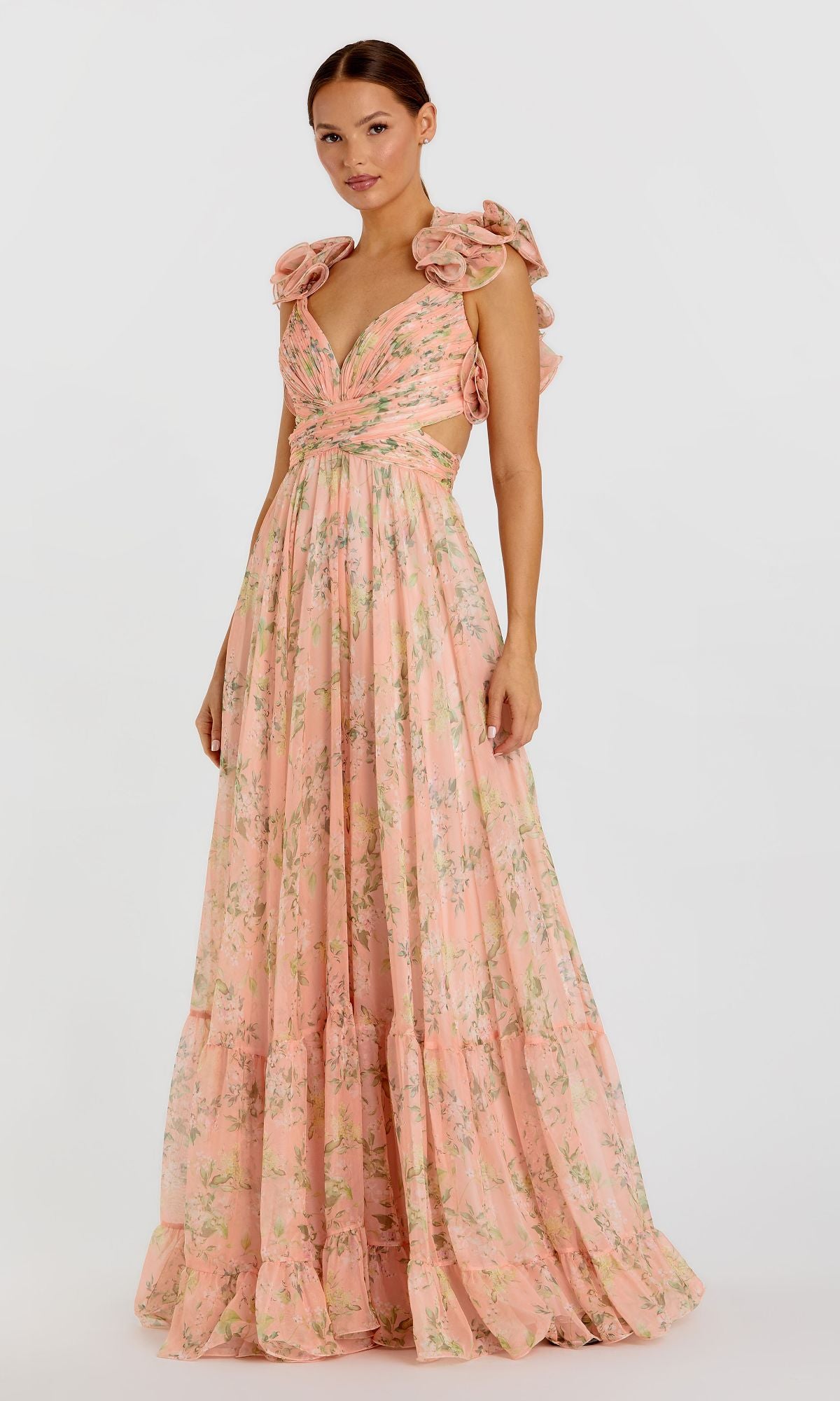 A woman models the Mac Duggal 67803 Ruffled Long Print Formal Dress, featuring a floor-length peach floral print, ruffled shoulders, and a deep V-neckline against a plain white background.