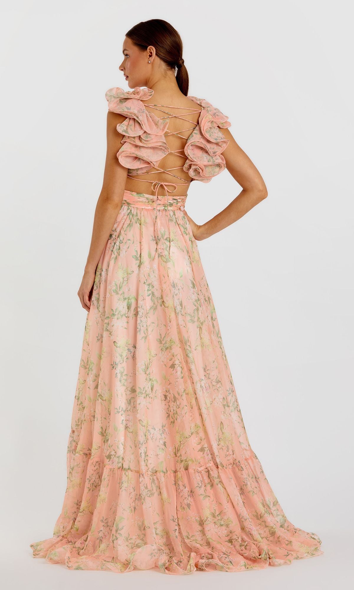 A woman models the Mac Duggal 67803 Ruffled Long Print Formal Dress, featuring a floor-length floral print, ruffled shoulders, and an open lace-up back, as she stands facing away from the camera.