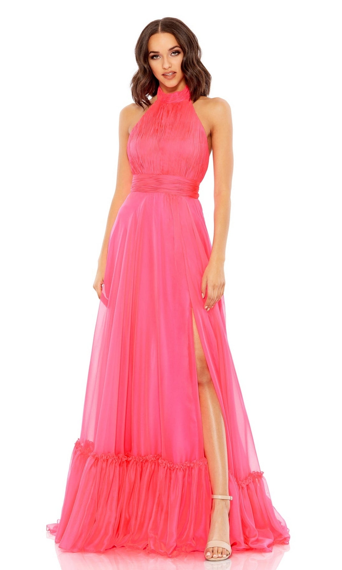 A woman wears the Mac Duggal 67816 Open-Back Halter Formal Dress: a sleeveless, floor-length bright pink gown with a high neckline, ruched bodice, and thigh-high slit, styled with strappy heels.