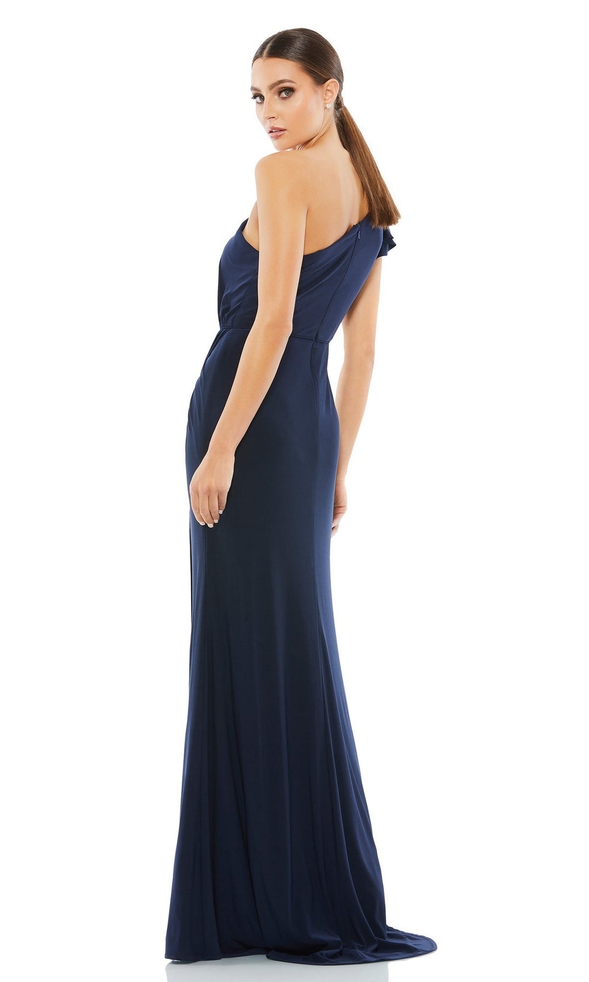 A woman with straight brown hair tied back models the Long Formal Dress 67858 by Mac Duggal, standing in a floor-length, one-shoulder gown with a subtle side slit, looking over her shoulder to showcase its elegant design.