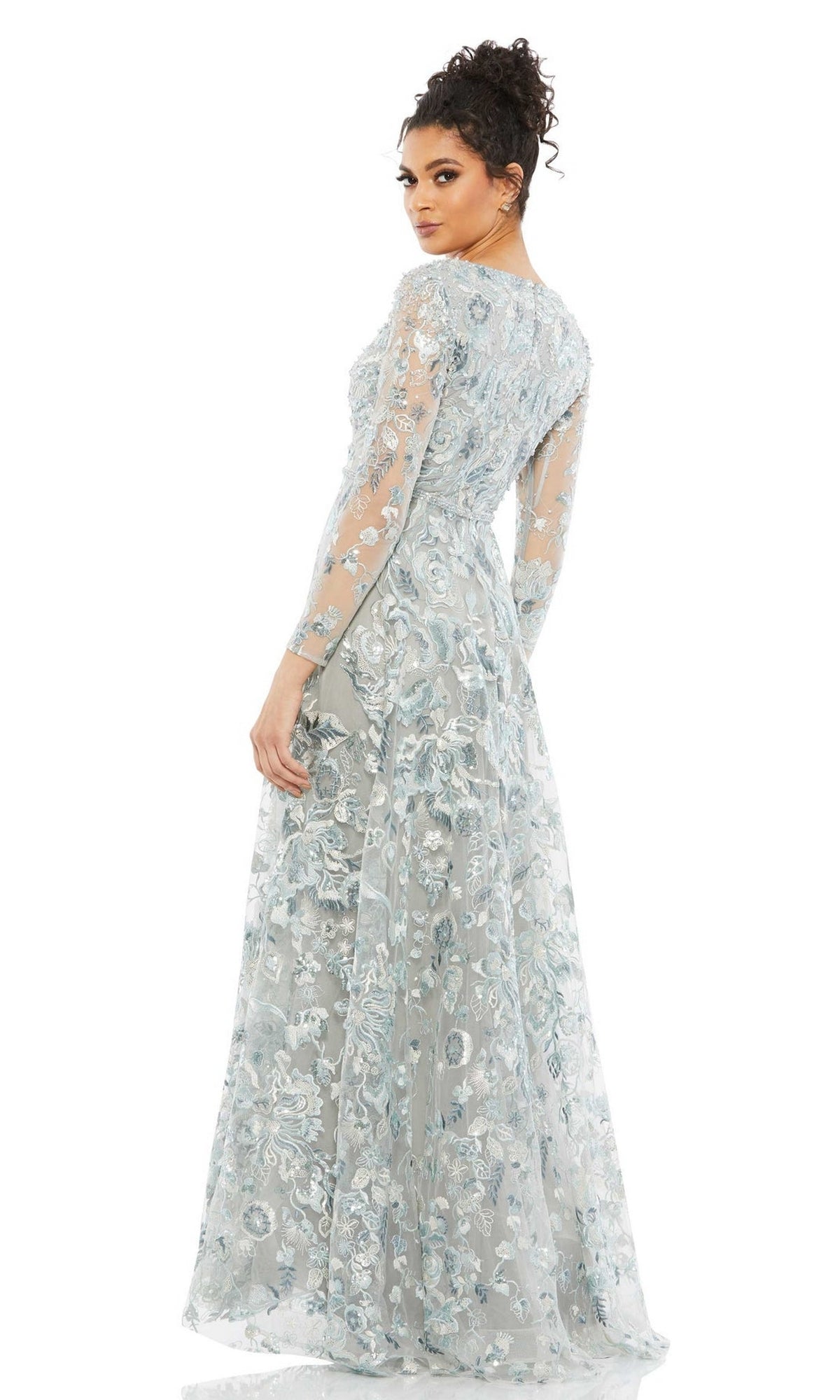 A woman models the Mac Duggal 67869 Long Sleeve Lace Formal Dress, a light blue floral embroidered evening gown with sheer long sleeves, as she stands looking over her shoulder against a plain white background.