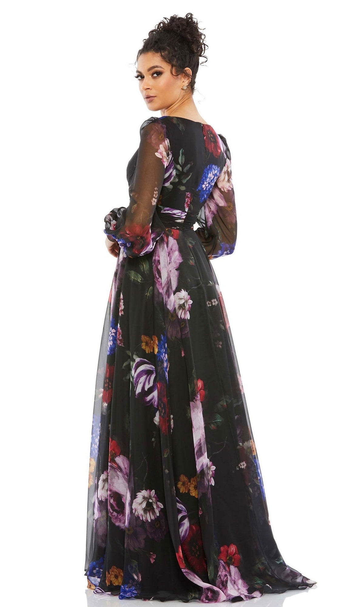 A woman is seen from the back, looking over her shoulder, wearing the Long Formal Dress 67872 by Mac Duggal, a long-sleeved, floor-length black floral gown with a colorful print and subtle empire waist.
