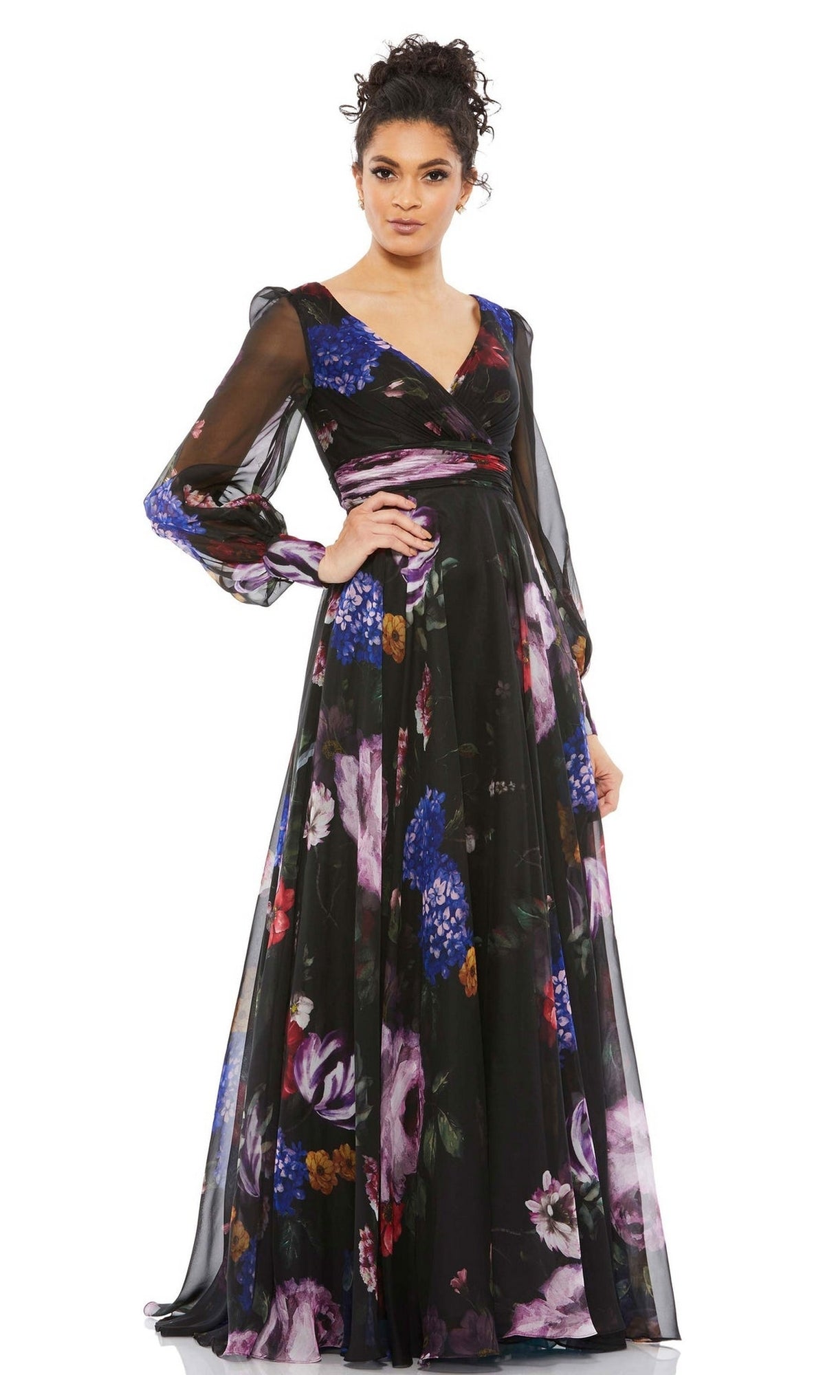 A woman models the Long Formal Dress 67872 by Mac Duggal, featuring sheer long sleeves, a colorful floral pattern, and an empire waist, posing with one hand on her hip against a plain white background.