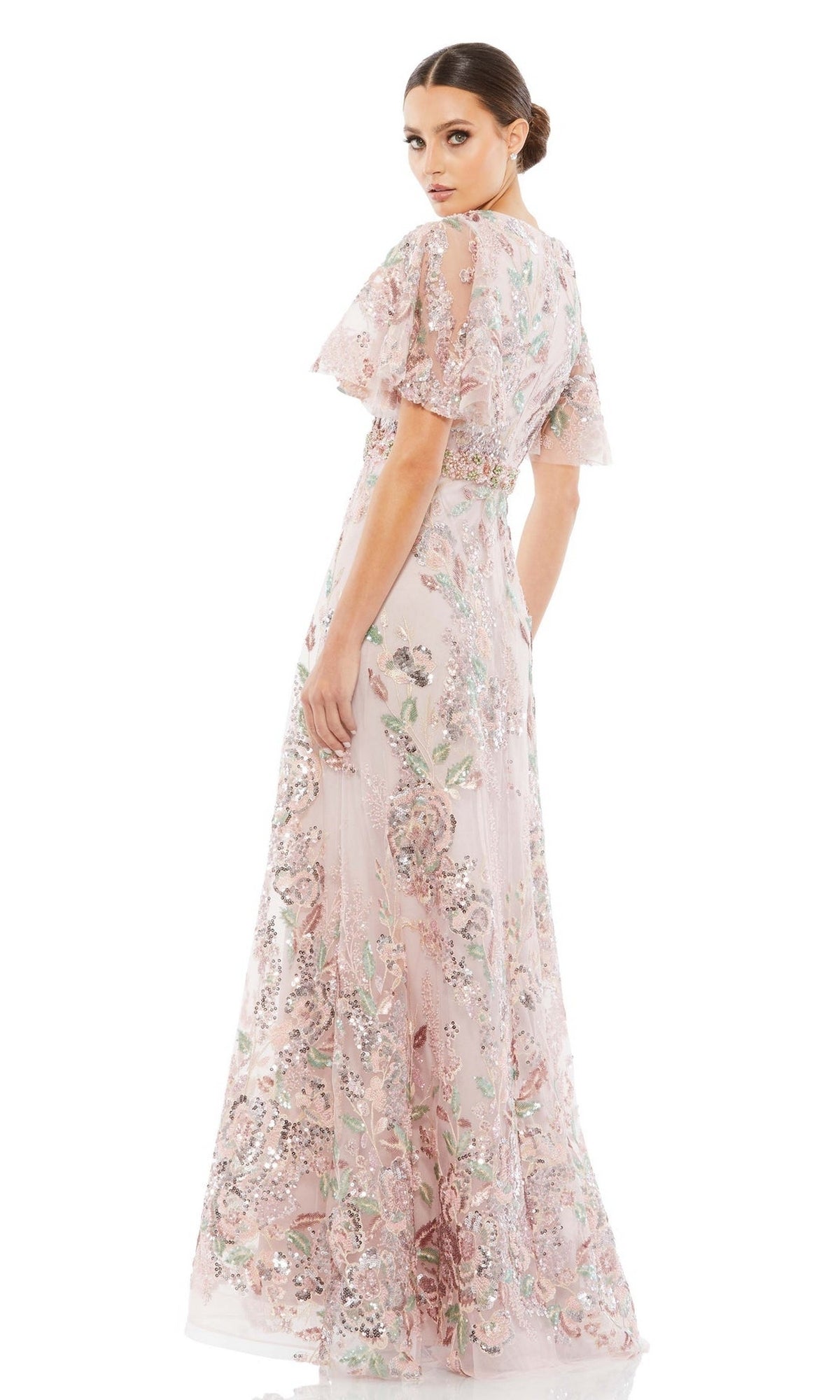 A woman models the Mac Duggal 67880 Short Sleeve Pink Formal Dress, a long floral-print gown with modest flutter sleeves, as she looks over her shoulder against a white background.