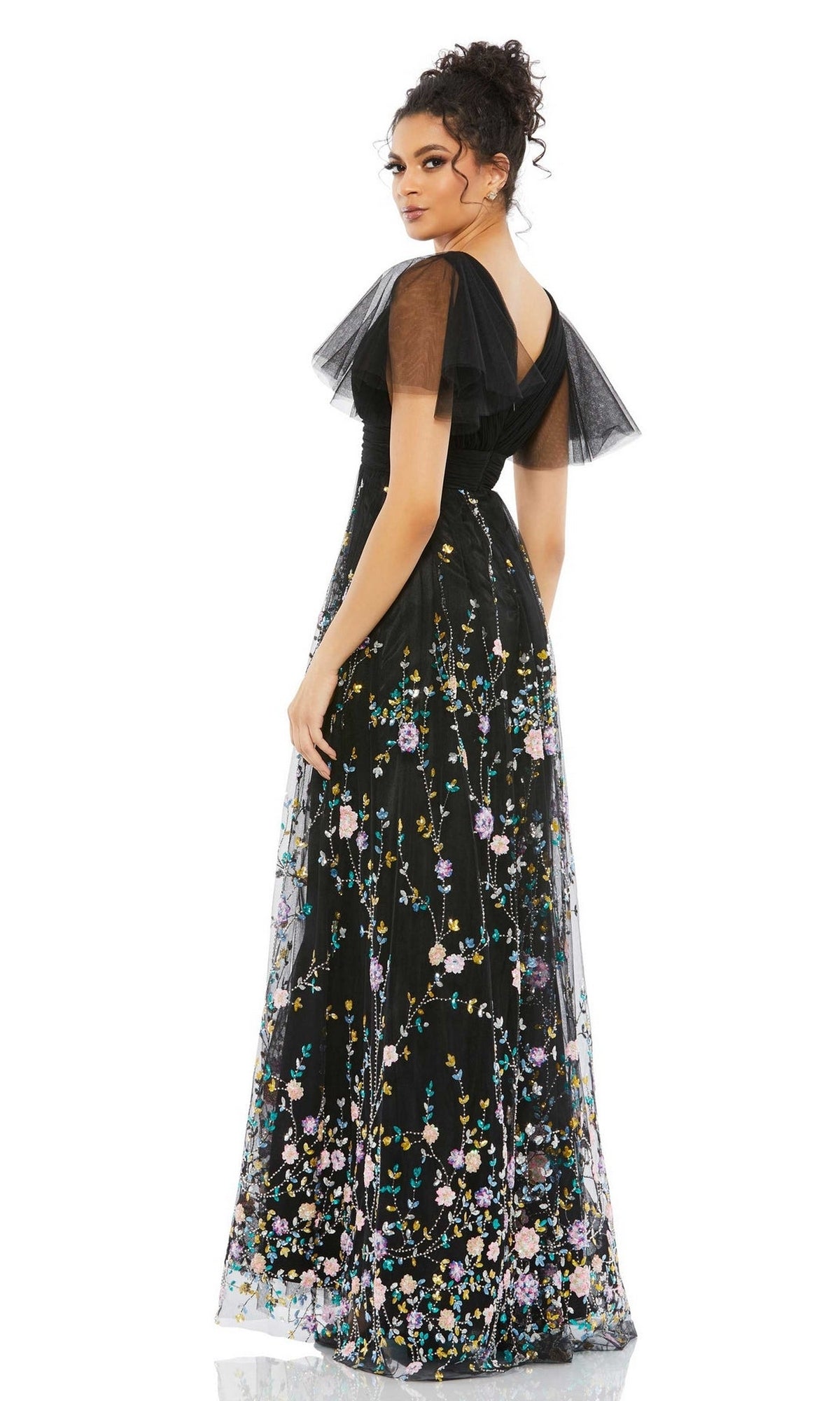 A woman faces away, wearing the Black Long Floral Formal Dress: Mac Duggal 67889, featuring sheer sleeves and colorful floral embroidery in classic Mac Duggal style, set against a white background.