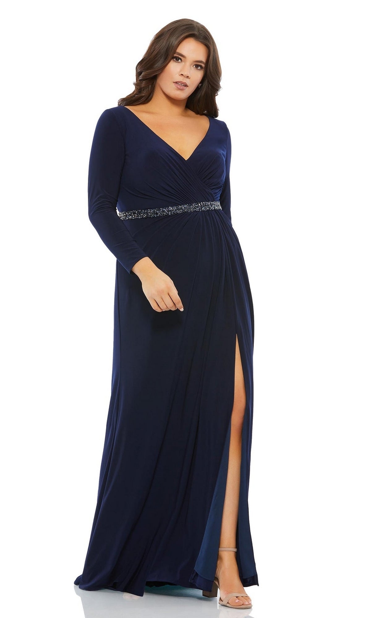 A woman stands confidently in heels, wearing the Long Plus-Size Prom Dress 67899 by Mac Duggal—a navy, floor-length gown with long sleeves, v-neckline, beaded waistband, and front slit.
