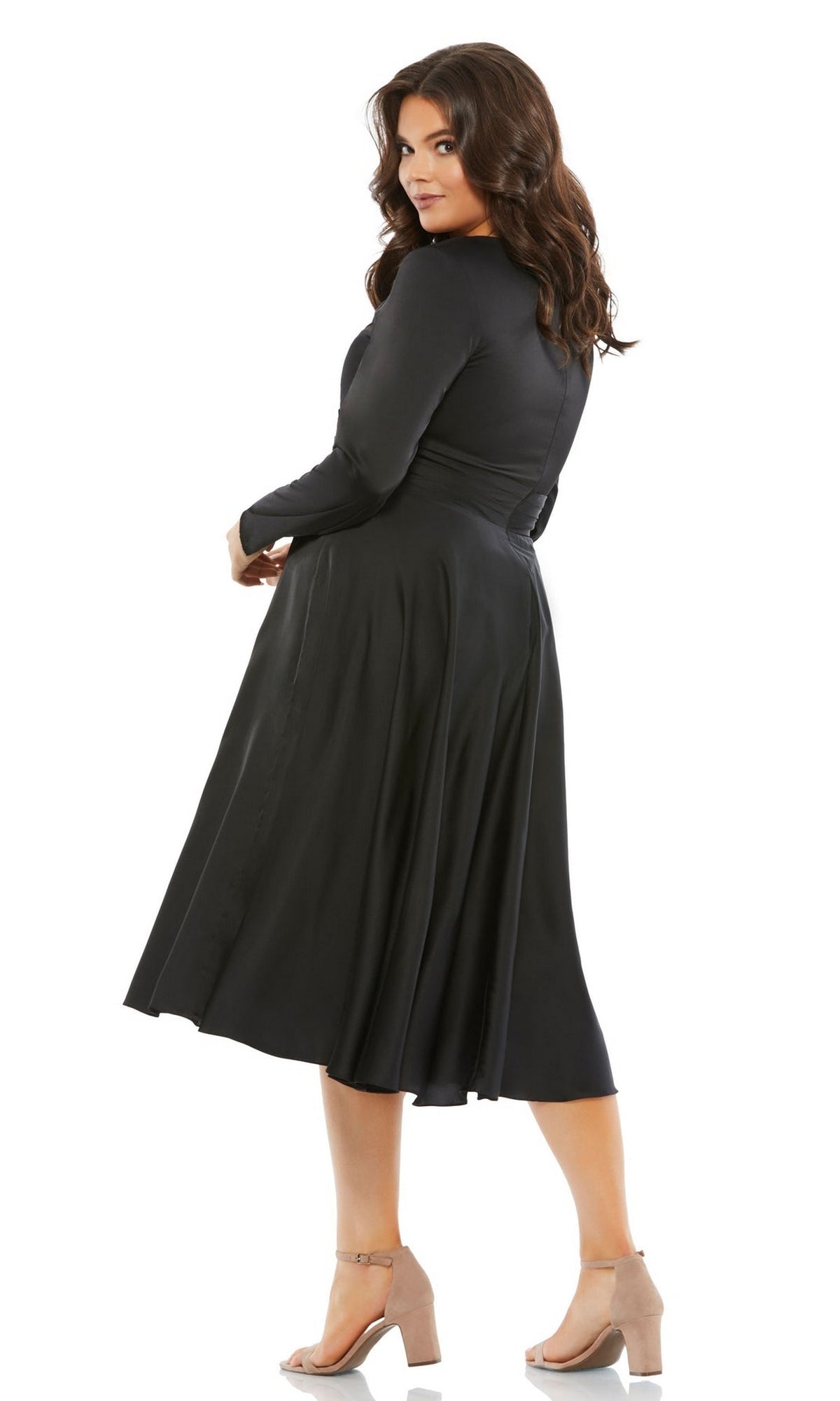 A woman models the Plus-Size Black Knee-Length Party Dress 67907 with long sleeves and beige heels, standing slightly turned and looking over her shoulder.