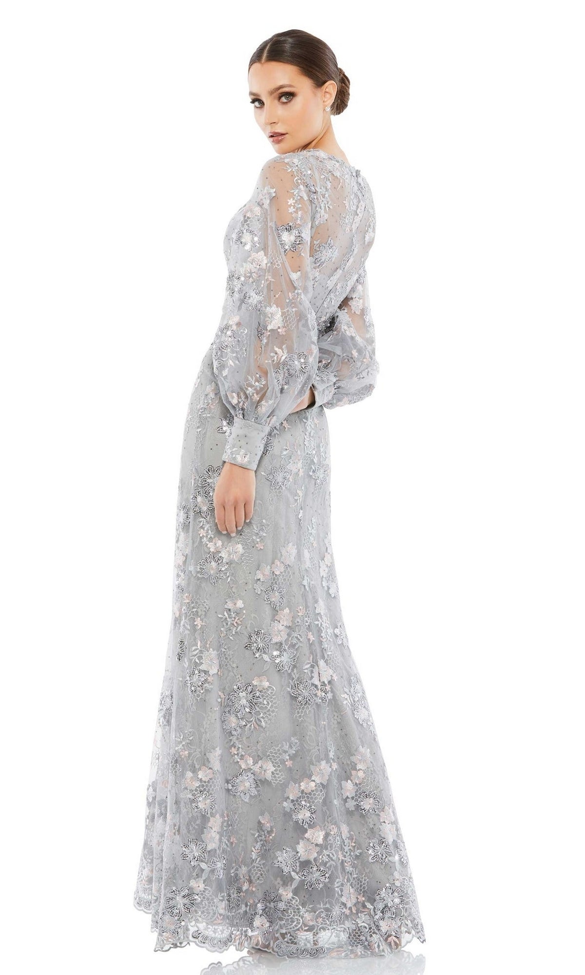A woman models the Long Formal Dress 67936 by Mac Duggal, featuring sheer light blue lace with floral embroidery, puffed sleeves, and a flowing floor-length skirt, posed against a white background.