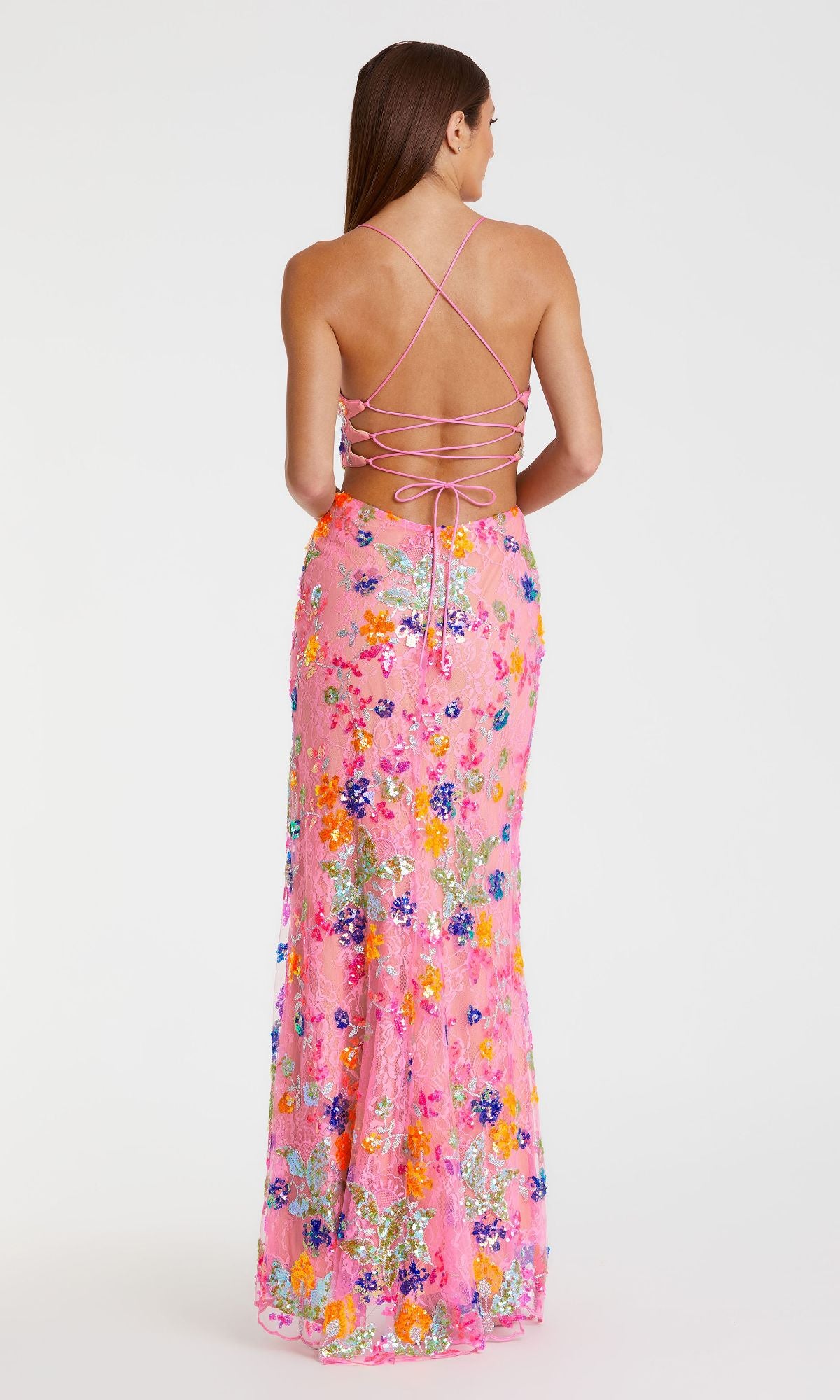 A woman stands with her back to the camera, wearing the Mac Duggal 68067 Sequin-Print Pink Formal Dress featuring crisscross straps and a long sequin floral skirt against a plain white background.