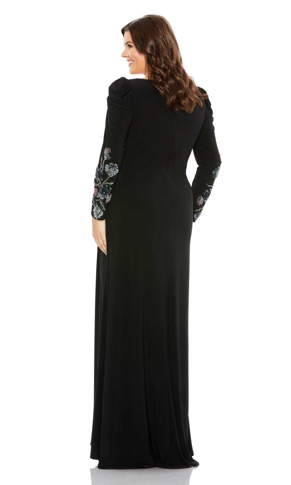 Woman models the Mac Duggal 68123 plus-size black formal dress, featuring floral embroidery on the sleeves, as she stands facing away and smiles over her shoulder against a plain white background.