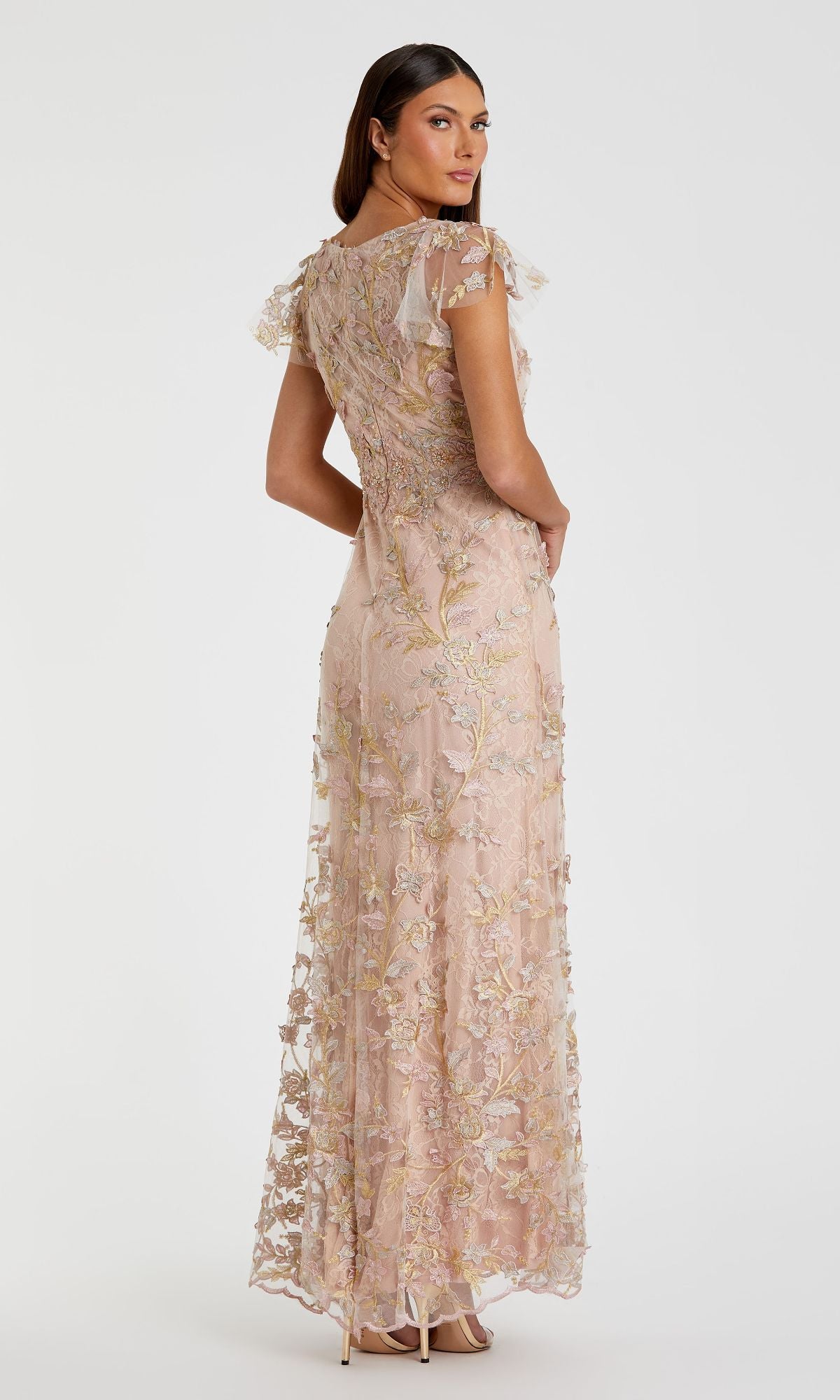 Woman in a blush Mac Duggal 68192 long beaded formal dress with lace floral embroidery and short sleeves, shown in a three-quarter back pose.