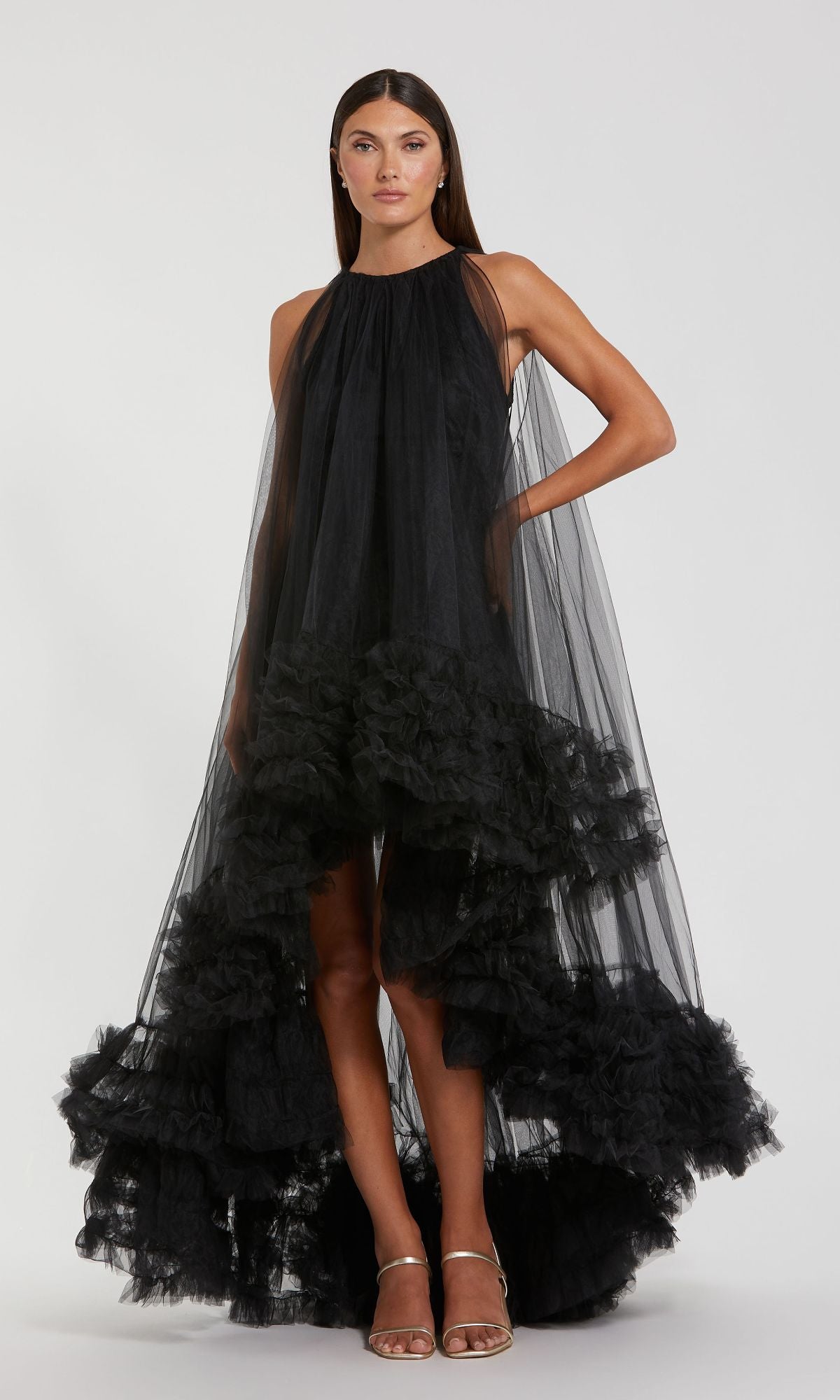 A woman models the Mac Duggal 68252 High-Low Trapeze Formal Dress—a sleeveless, high-neck black tulle gown with a ruffled high-low hem—against a plain background.