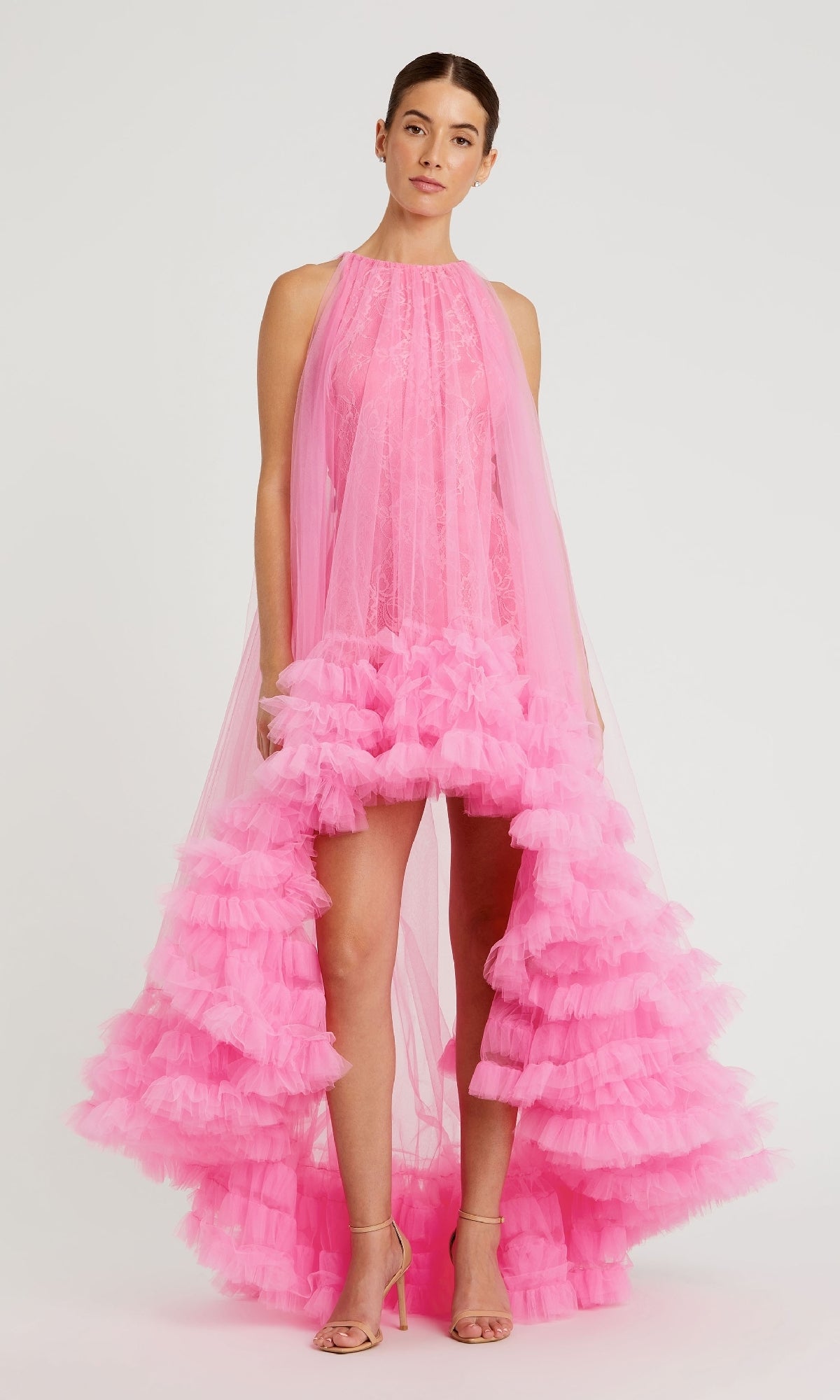 A person stands against a plain background wearing the Mac Duggal 68252 High-Low Trapeze Formal Dress in bright pink with ruffles and lace details, paired with nude heeled sandals.