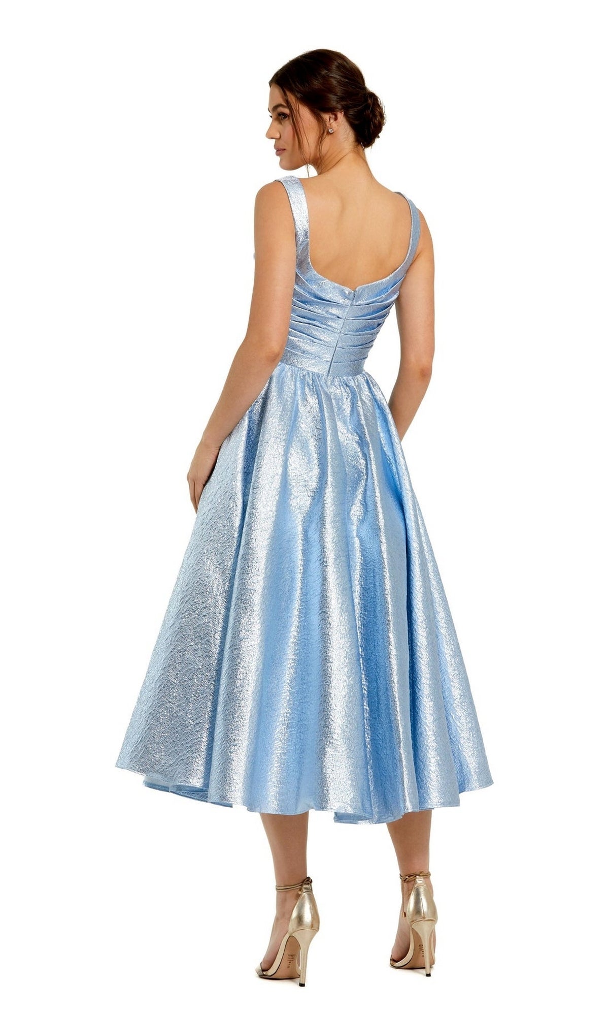 A woman stands facing away in the Mac Duggal 68350 tea-length wedding-guest dress—light blue, sleeveless, with a fitted bodice and flared skirt—paired with metallic high heels.