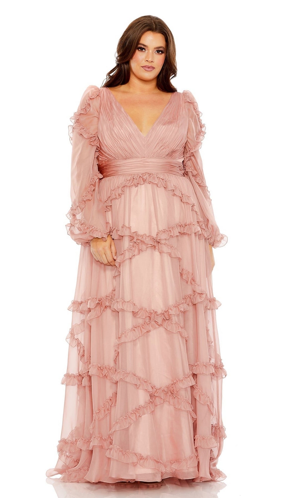A woman models the Long Plus-Size Prom Dress 68425 by Mac Duggal—blush pink, floor-length, with ruffled details, long sleeves, and a deep V-neckline—against a plain white background.