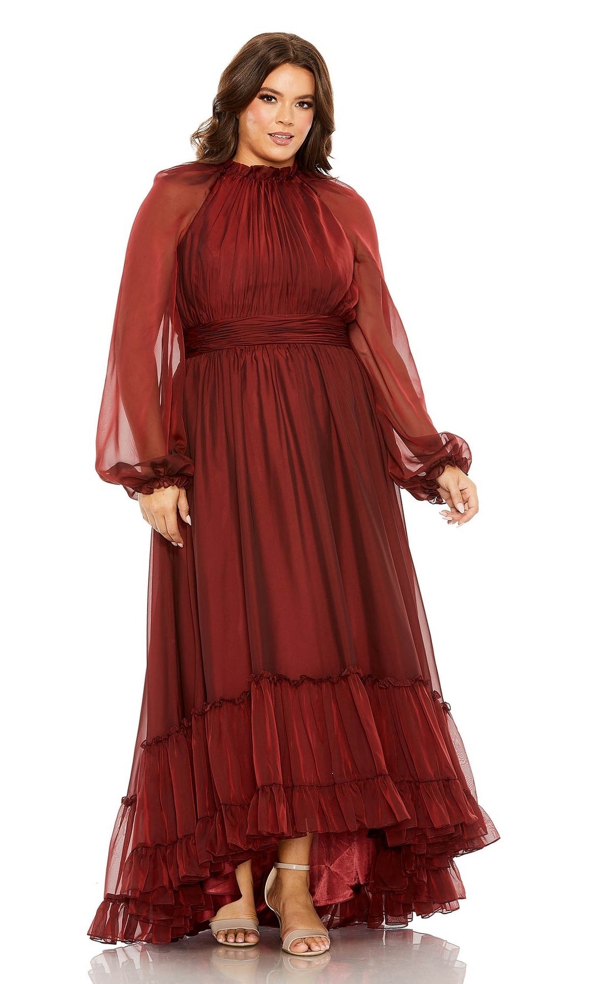A woman stands and looks at the camera wearing the Long Plus-Size Prom Dress 68434 by Mac Duggal, featuring a burgundy hue, sheer sleeves, ruffle hem, and a layered skirt.