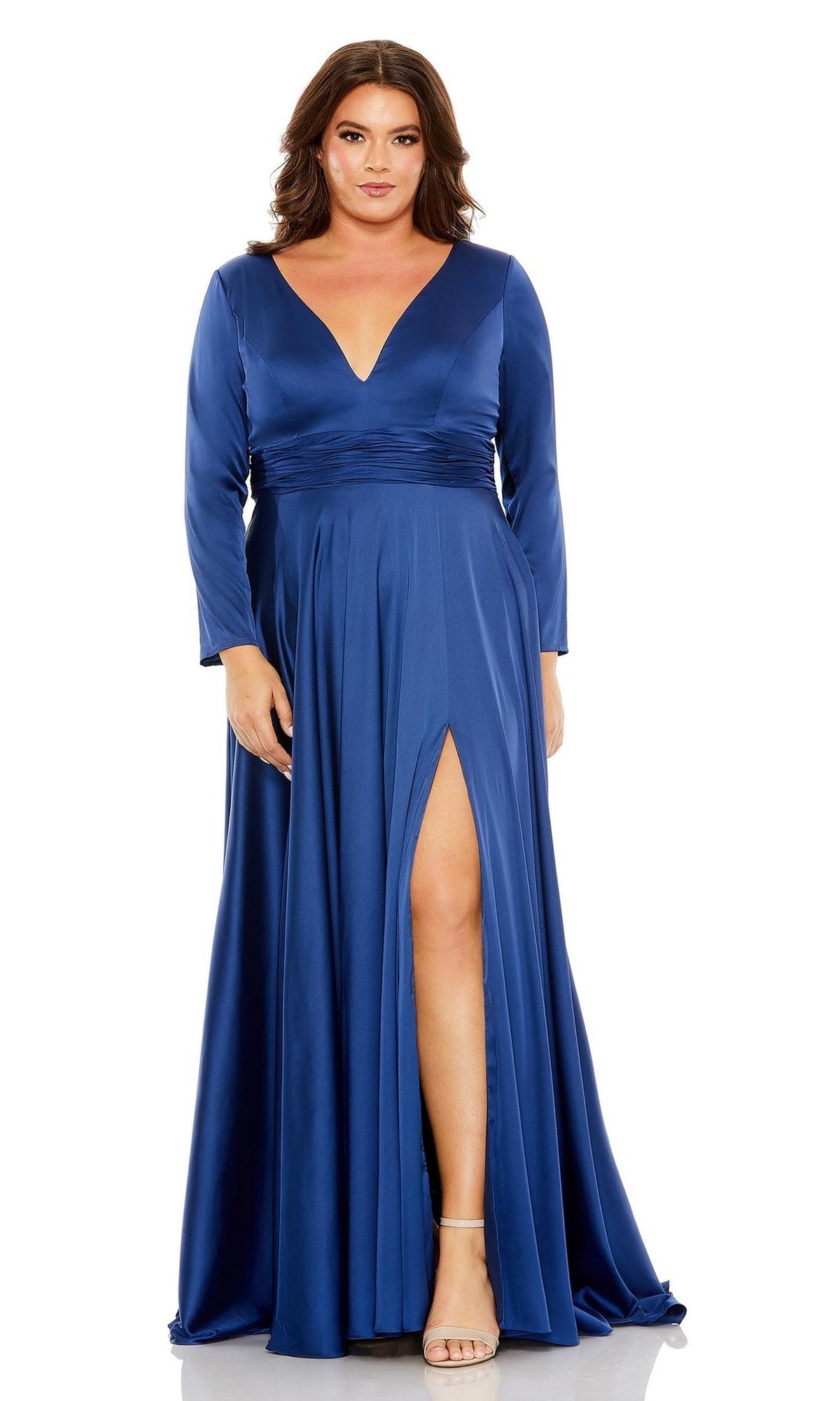 A woman models the Mac Duggal 68436 plus-size long sleeve prom gown in deep blue satin, featuring a deep V-neckline and thigh-high front slit, against a white background.