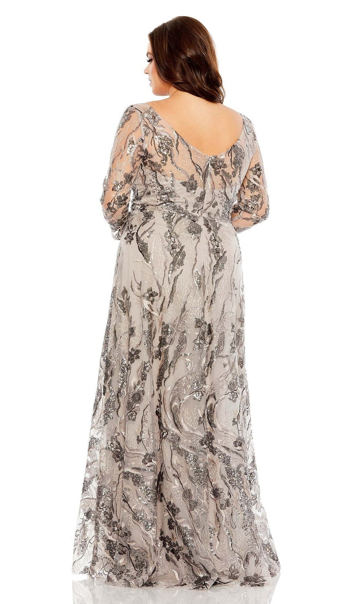 A woman faces away, showcasing the low V-back and floor-length hem of the Long Plus-Size Prom Dress 68439 by Mac Duggal—a sheer, silver-grey A-line gown with intricate embroidery.