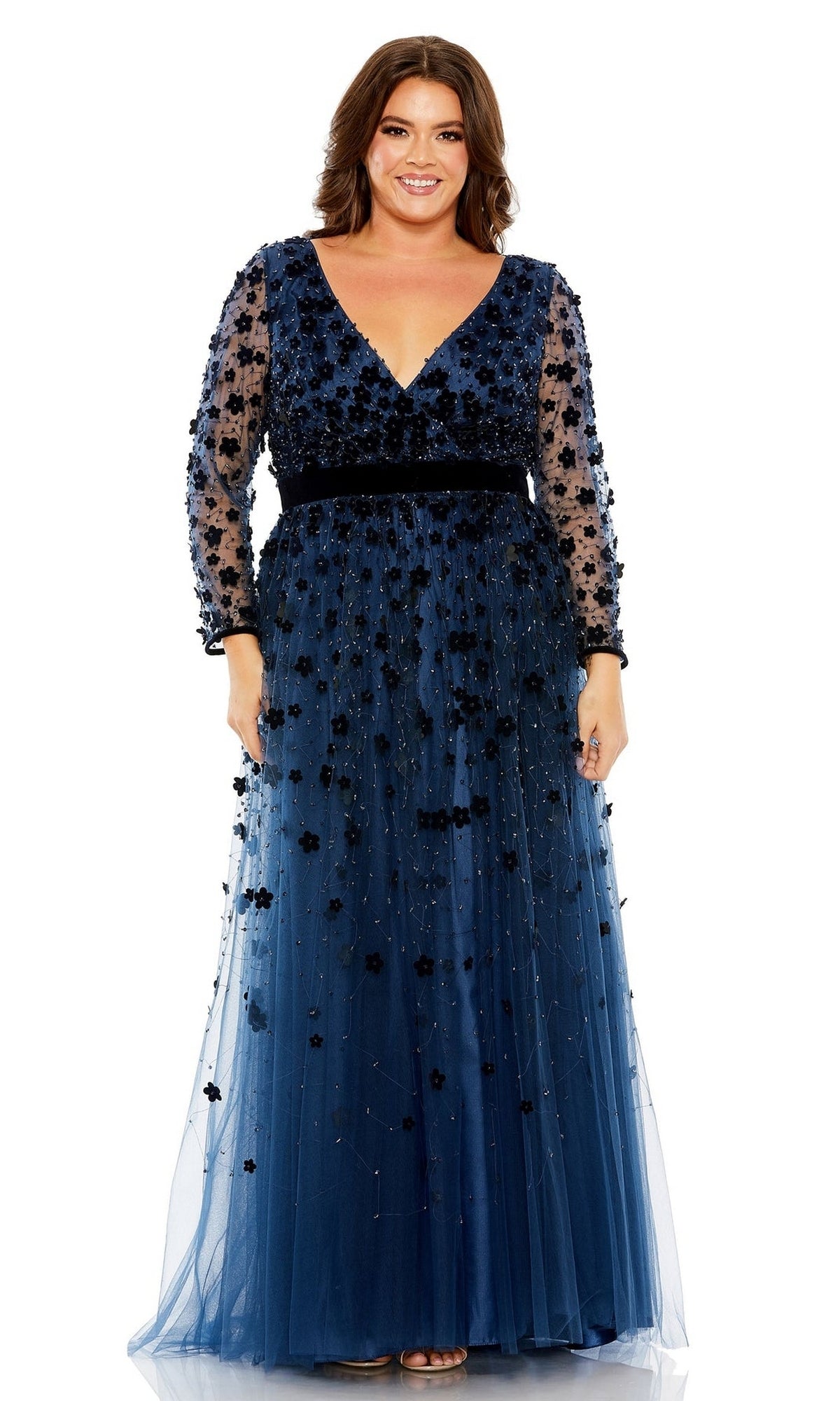 A woman stands smiling in the Long Plus-Size Prom Dress 68440 by Mac Duggal, a navy blue floor-length gown with floral embellishments and a tulle overlay.