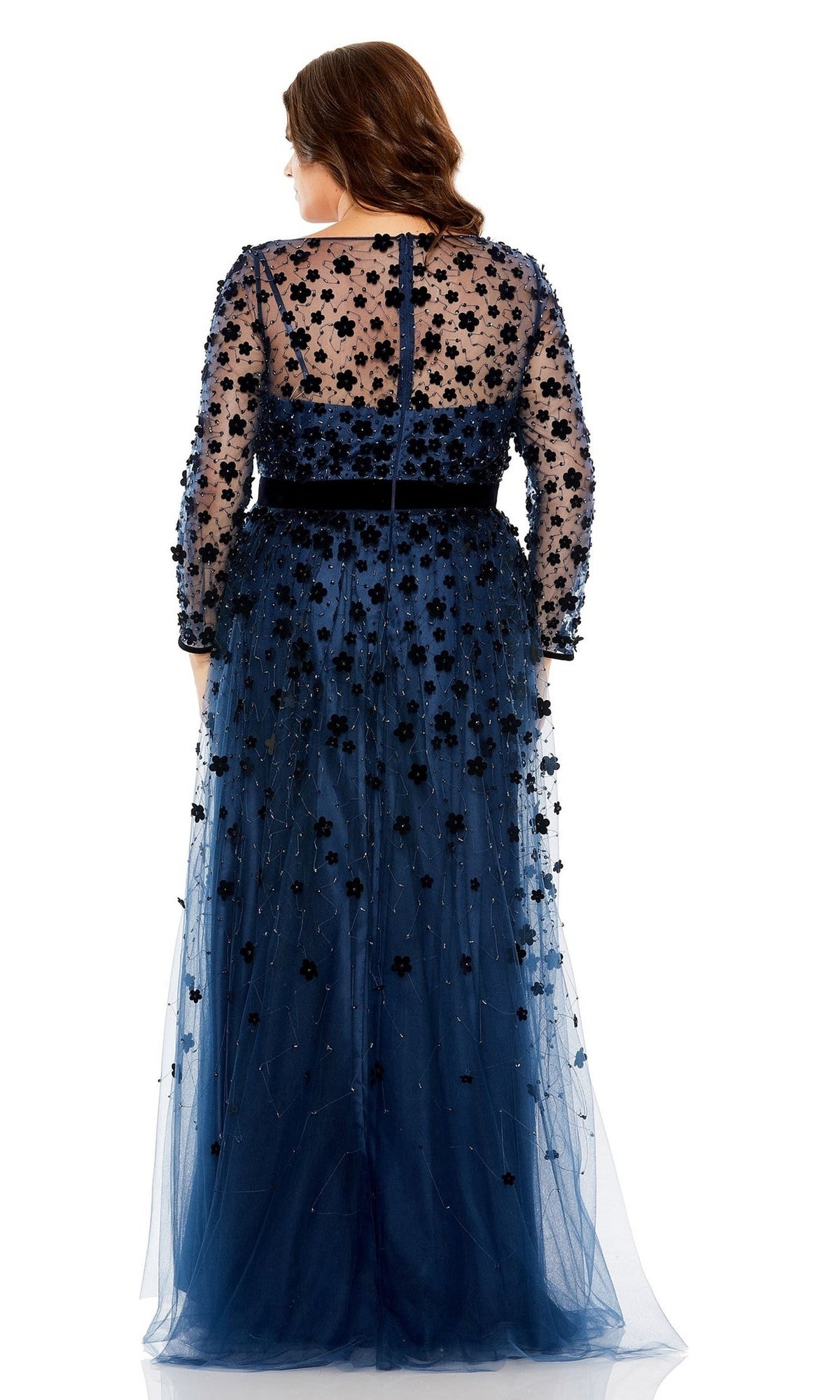 A woman is seen from the back wearing the Mac Duggal Long Plus-Size Prom Dress 68440, a formal floor-length navy gown with sheer sleeves and bodice, adorned with black floral appliqué.