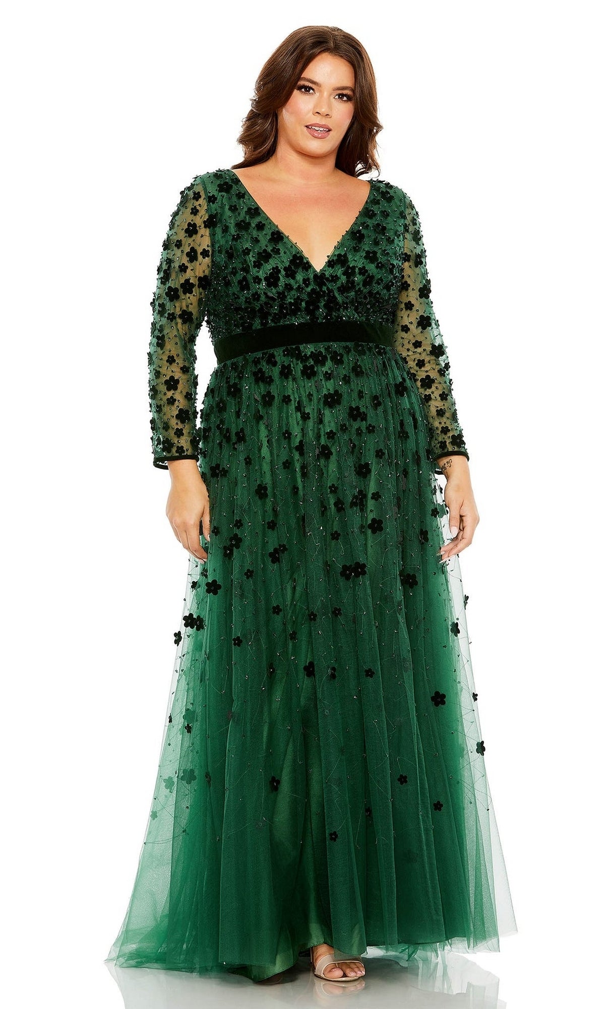 A woman stands against a white background wearing the Long Plus-Size Prom Dress 68440 by Mac Duggal, featuring floor-length deep green tulle, sheer sleeves, and black floral embellishments.