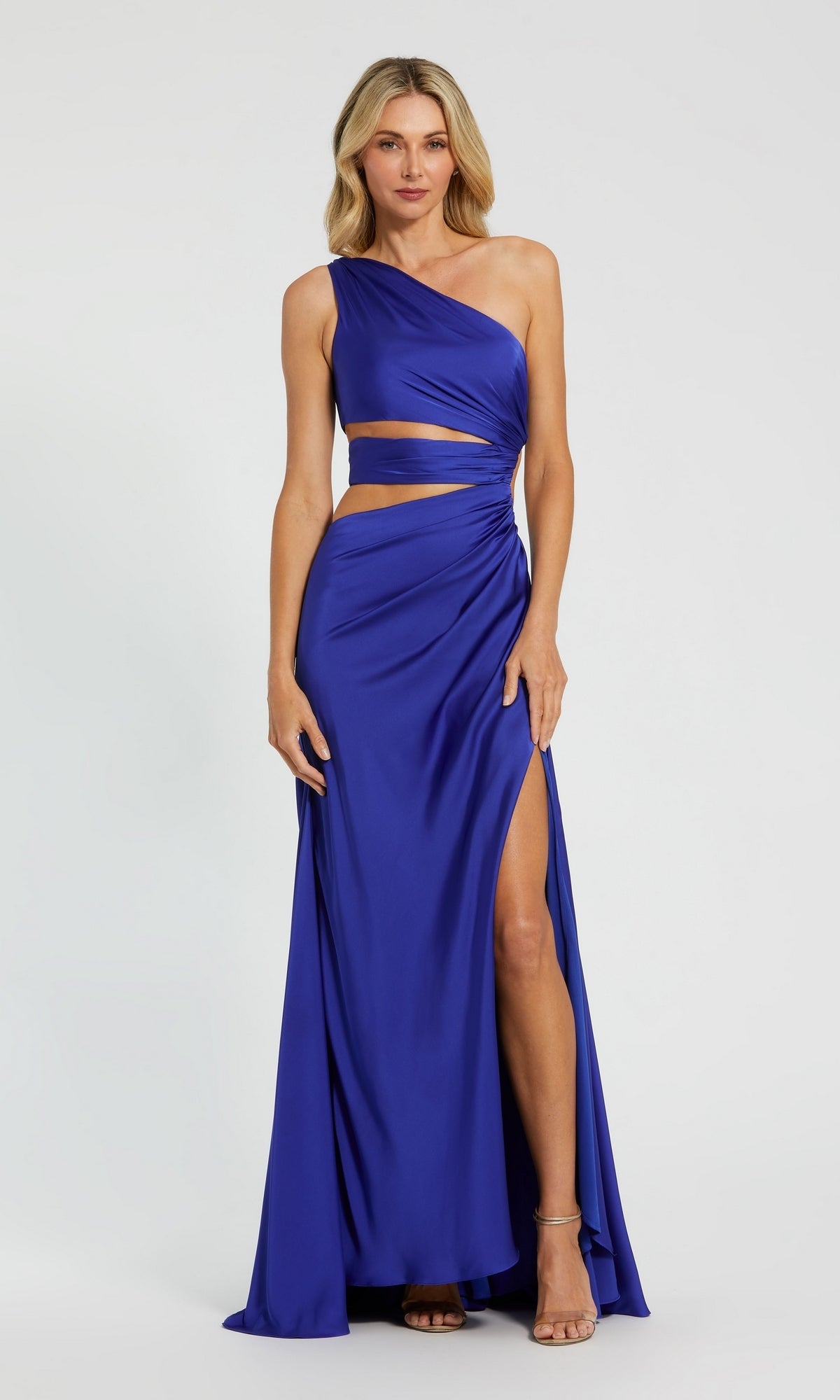 A woman models the Mac Duggal 68485 One-Shoulder Cut-Out Formal Gown in royal blue, featuring a high slit and waist cut-outs, against a plain background.