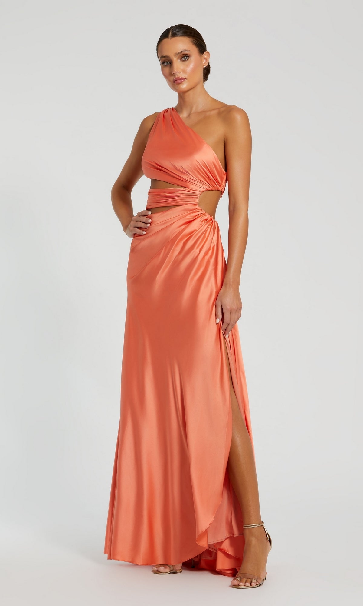 A woman wears the Mac Duggal 68485 One-Shoulder Cut-Out Formal Gown in coral-orange satin, featuring side cutouts and a thigh-high slit, styled with high-heeled sandals against a plain background.