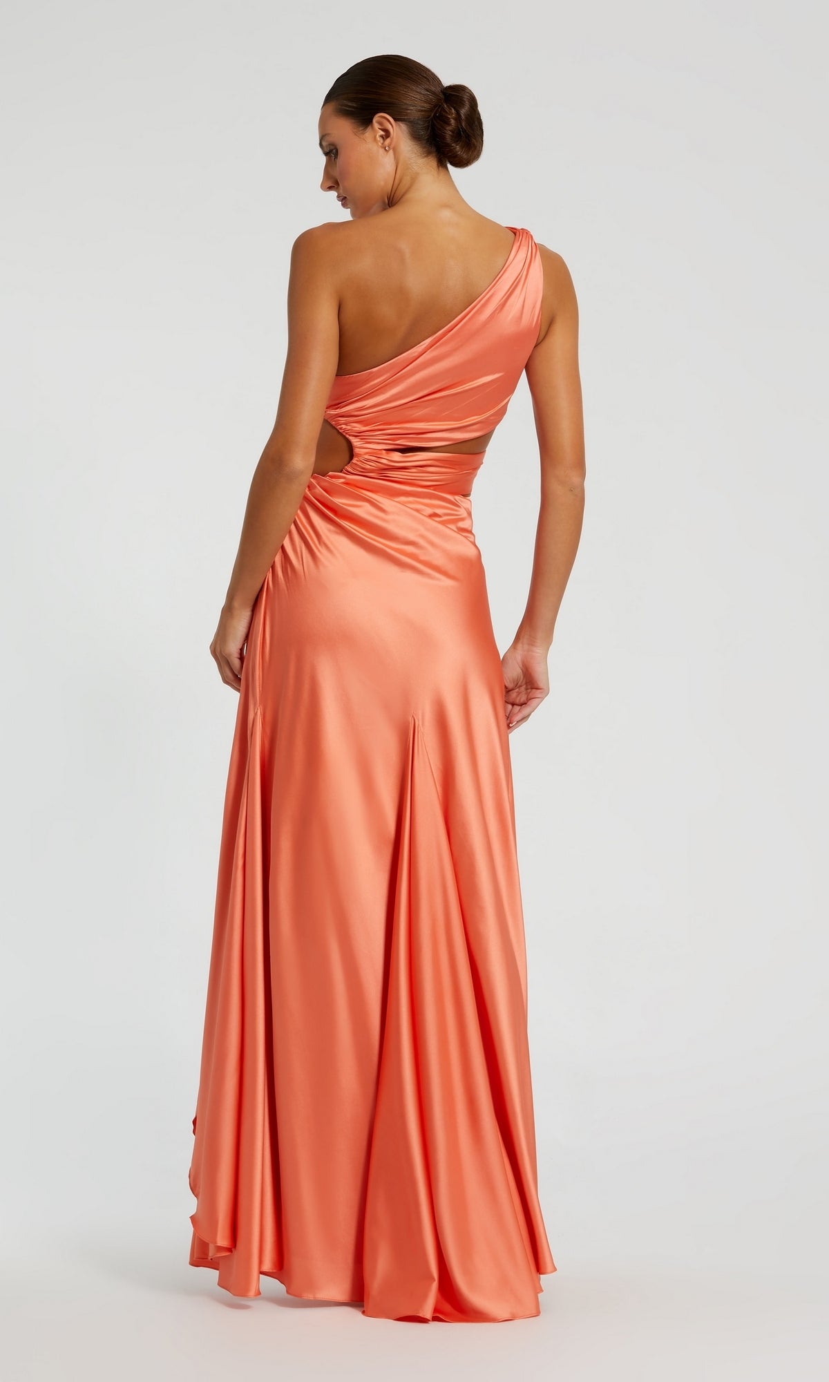 A woman in the Mac Duggal 68485 One-Shoulder Cut-Out Formal Gown, shiny coral with a waist cutout, stands facing away with her hair in a low bun against a plain light background.