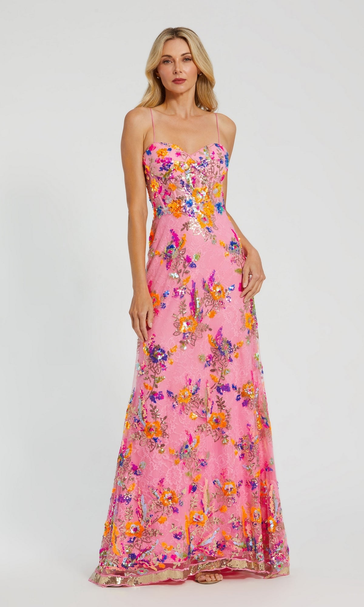 A woman stands against a plain background wearing the Long Formal Dress 68508 by Mac Duggal—a floor-length pink lace gown with colorful floral patterns and a sweetheart neckline.