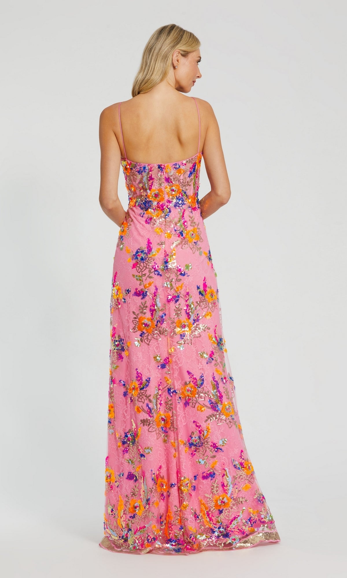 A woman stands facing away, wearing the Long Formal Dress 68508 by Mac Duggal—a long pink lace dress with thin straps and colorful sequin floral patterns.