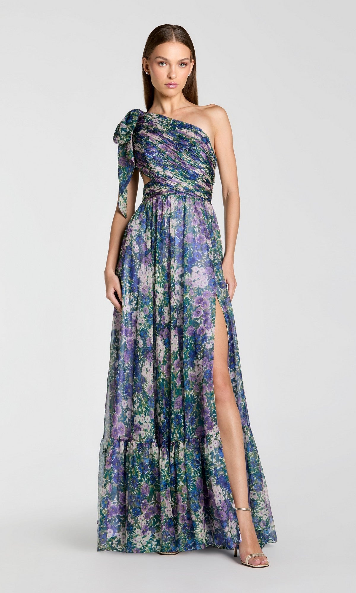 A woman poses against a plain background in the Mac Duggal 68553, a one-shoulder floral formal gown with a thigh-high slit, embodying modern elegance.