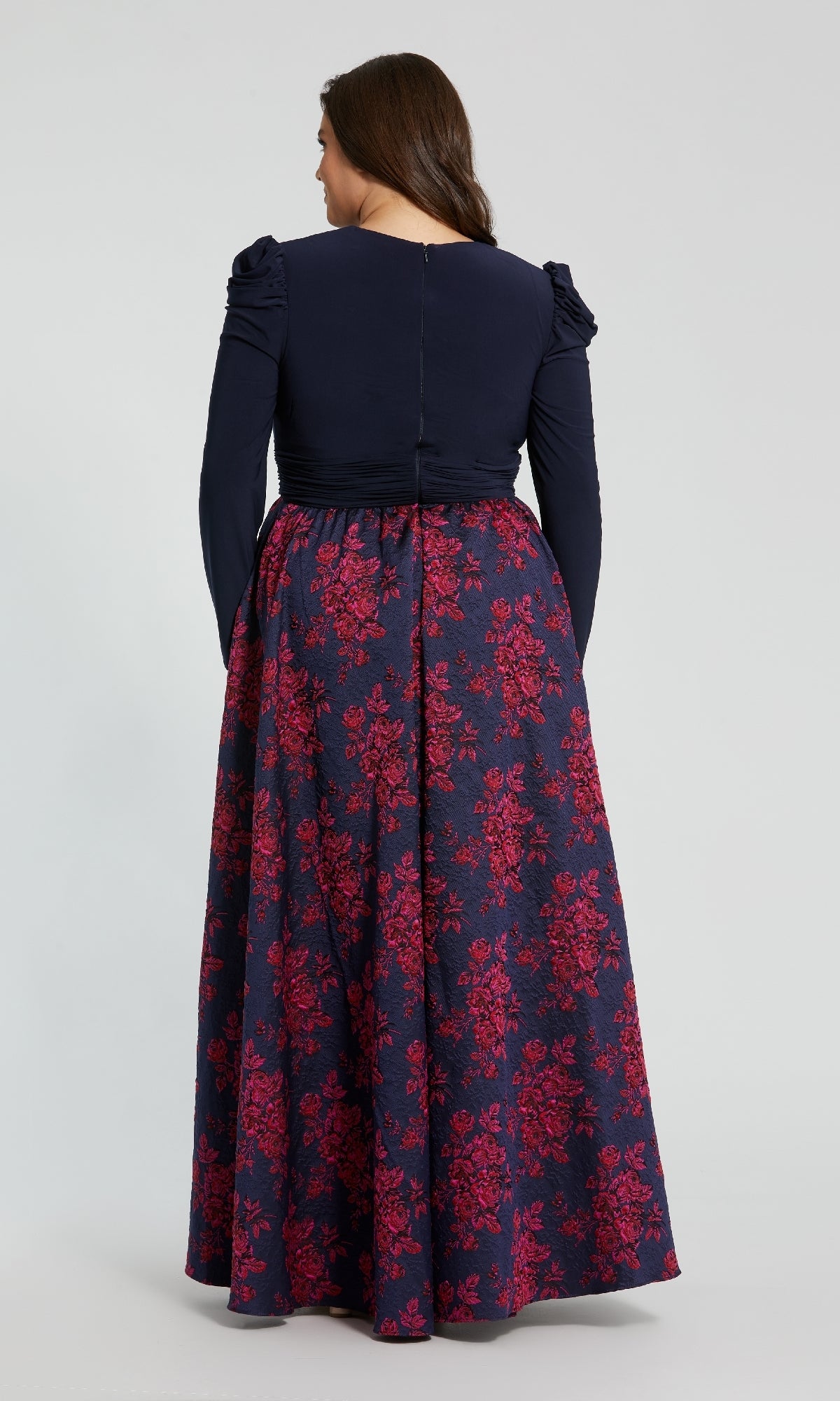 A woman stands facing away, wearing the Mac Duggal 68556 plus-size long blue formal dress with navy sleeves and a dark blue skirt featuring a red floral pattern.
