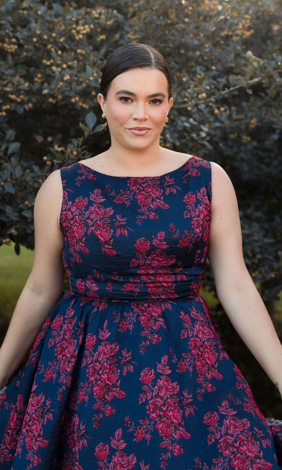 A woman stands outdoors before green foliage, wearing the Plus-Size Knee-Length Print Party Dress 68557—a sleeveless floral navy blue dress with pink patterns.