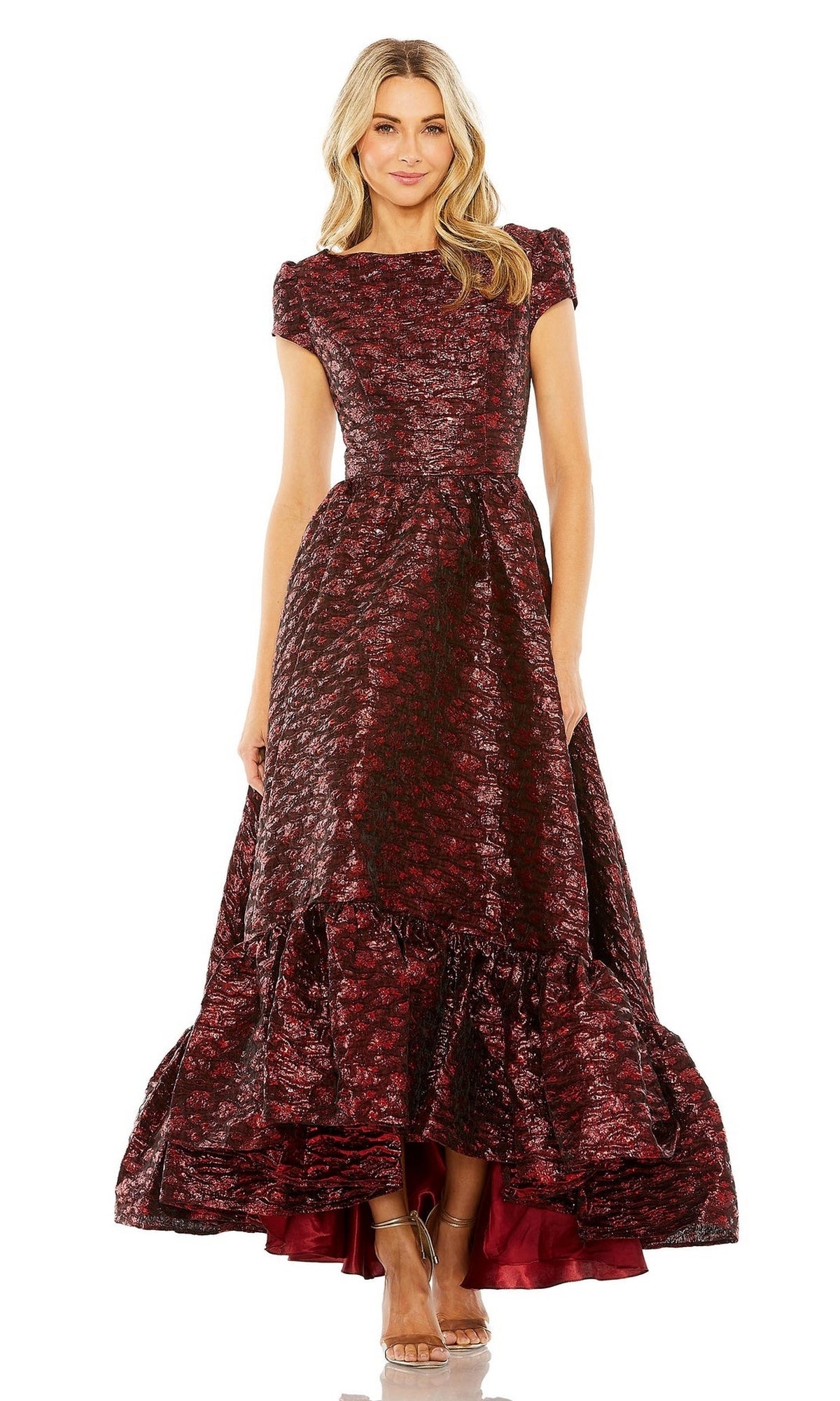 A woman wears the Long Formal Dress 68623 by Mac Duggal, a floor-length burgundy and black brocade gown with short sleeves, ruffled hem, and high-low skirt, paired with clear high-heeled shoes.